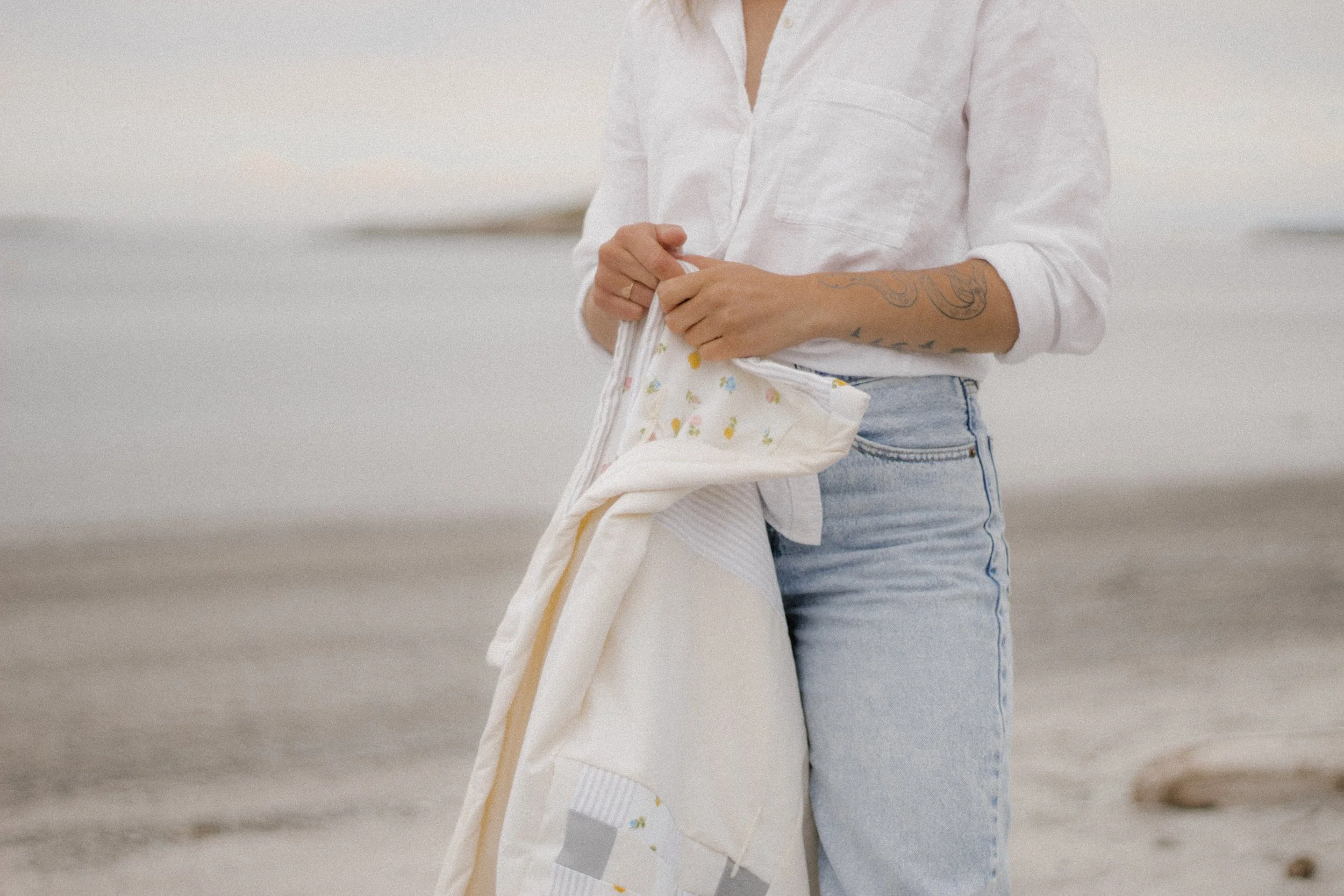 Person with tattoo holding a patterned quilt on a beach, wearing a white shirt and jeans.