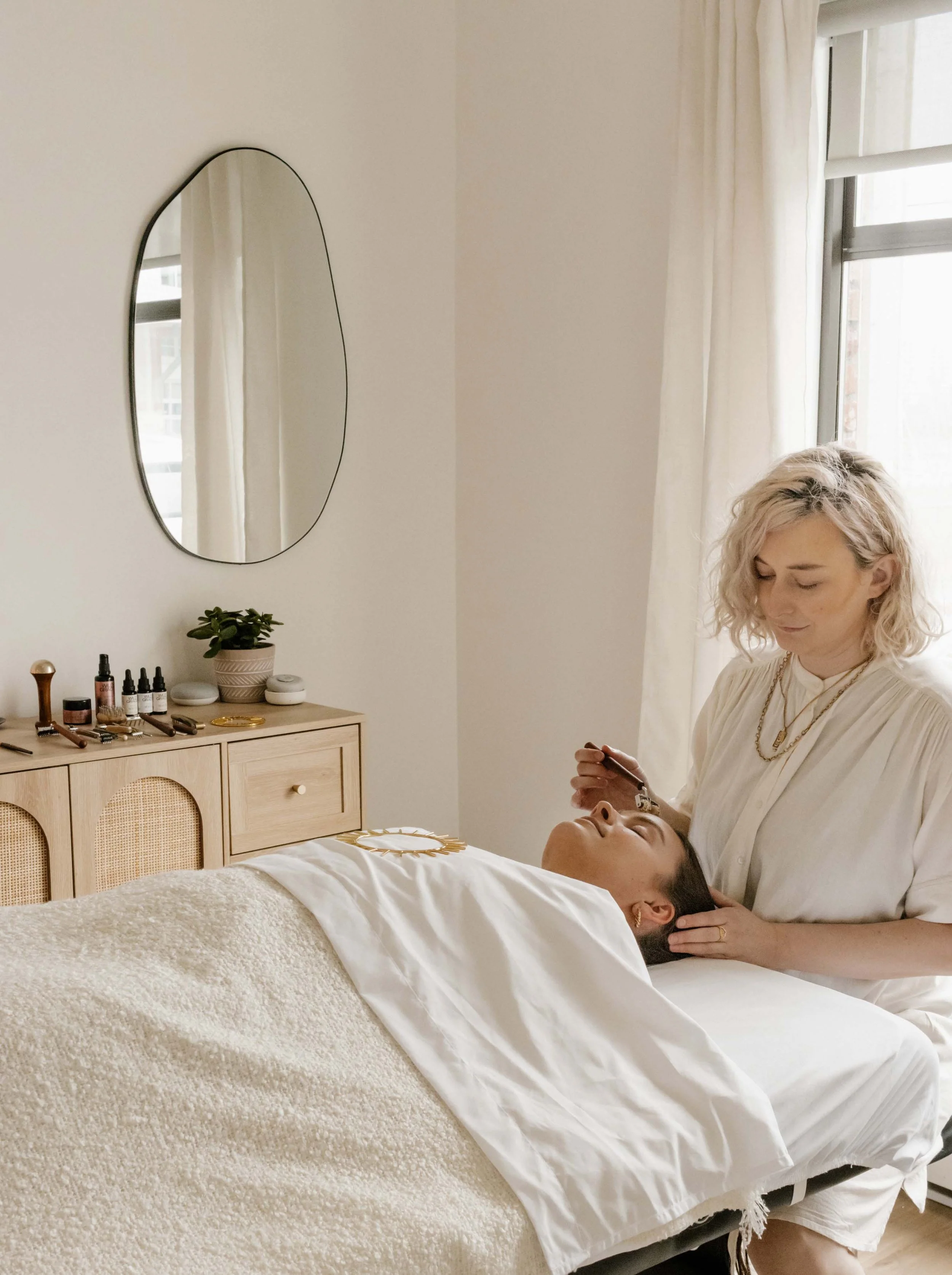 A person receiving a facial treatment in a spa room with a therapist using a roller on their face. The room has a minimalist decor with a mirror, wooden cabinet, and various skincare products.