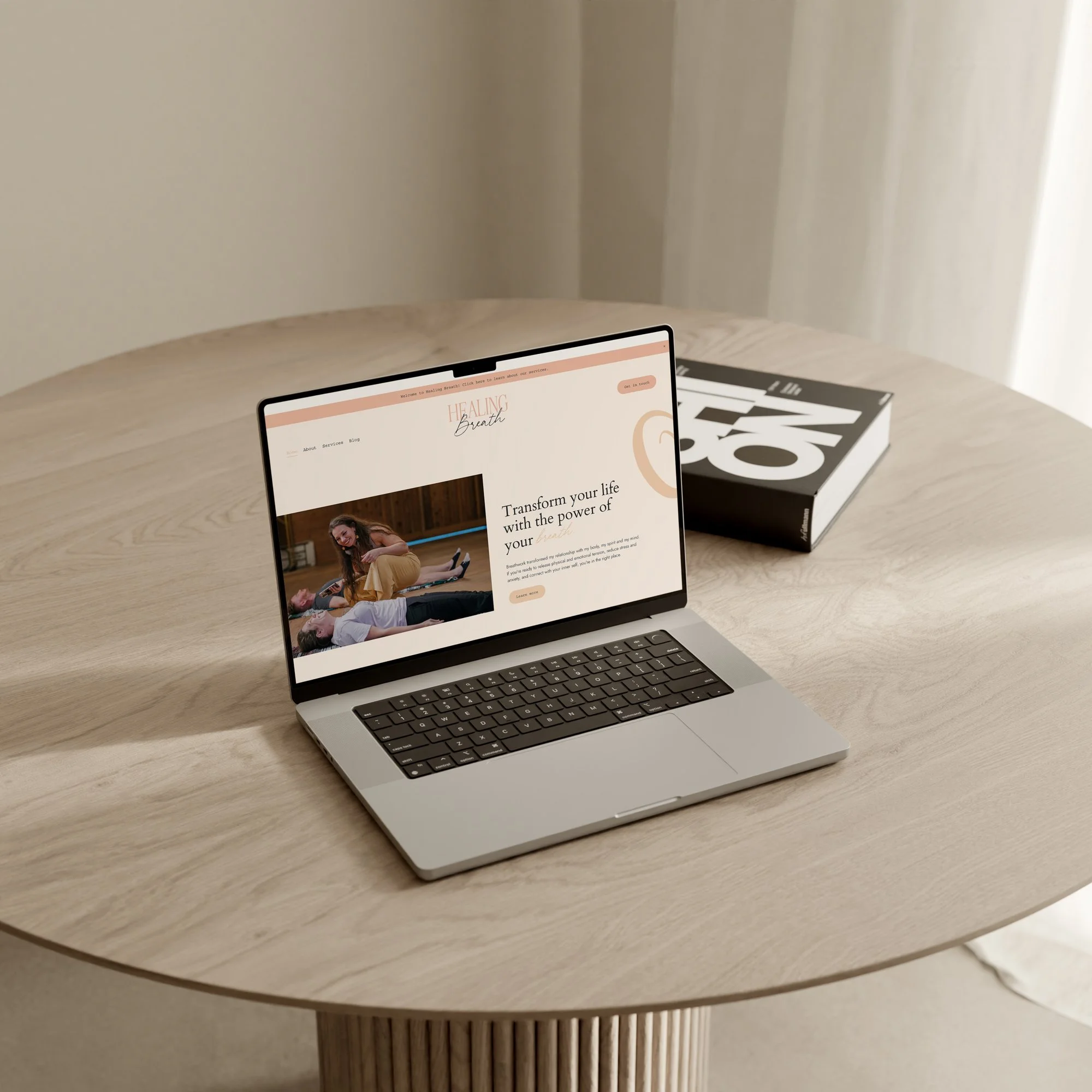 A laptop on a wooden round table displaying a website titled "Healing Breath" with a picture of people practicing yoga, and a book with large black print is placed beside the laptop.