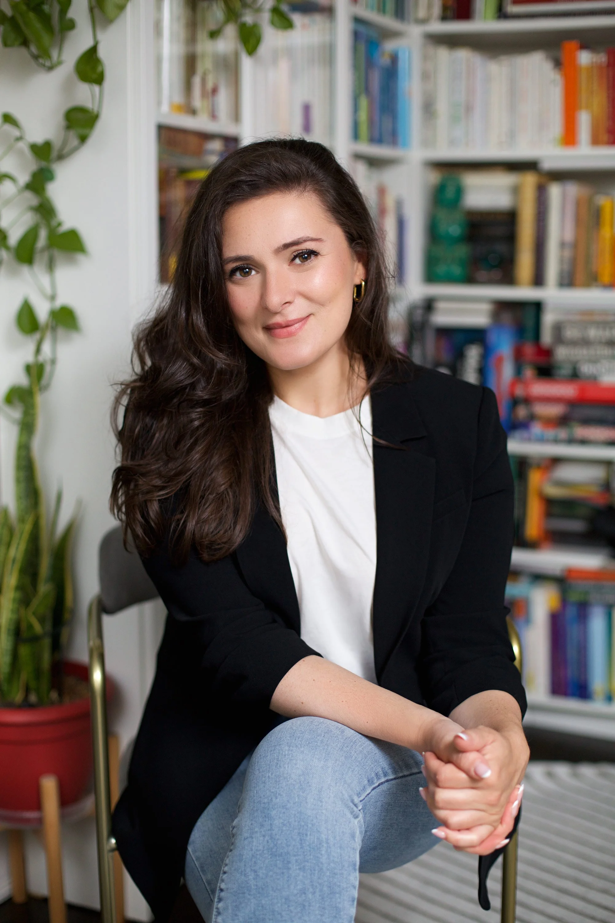A woman with long brown hair, wearing a black blazer and white t-shirt, sitting in front of a bookshelf filled with colorful books and a green plant in a red pot.