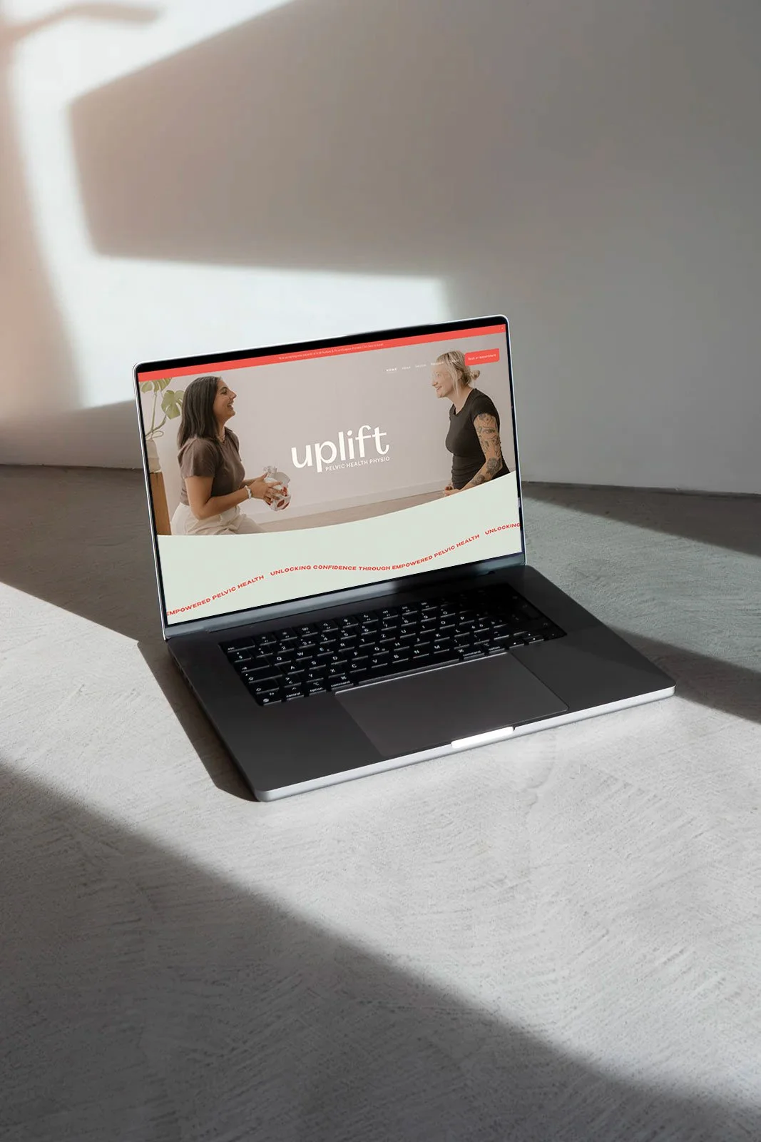 Laptop on a light-colored surface displaying a website for 'uplift,' featuring two women smiling and talking, with a plant in the background on the website.