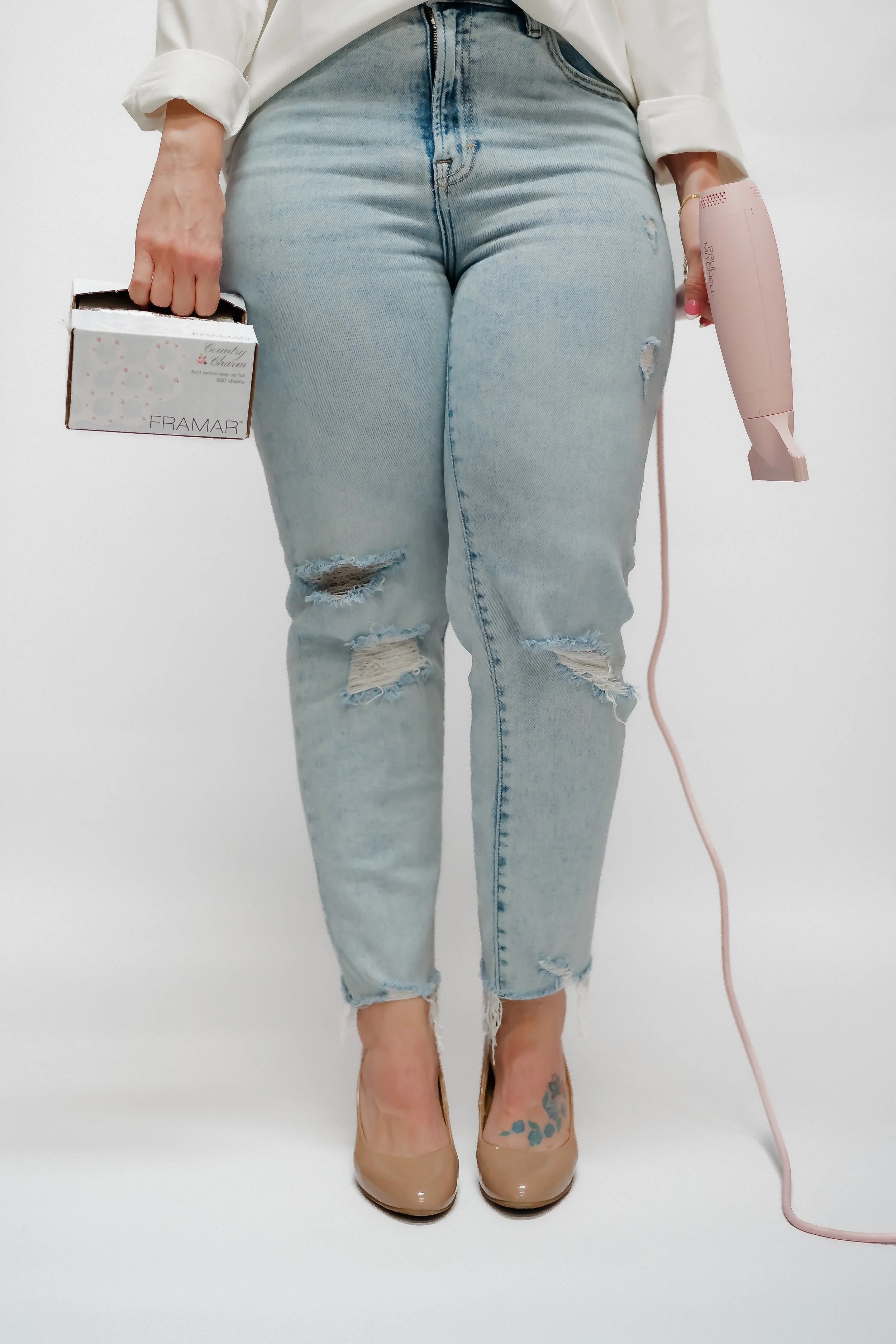 Close-up of a woman wearing light-wash ripped jeans, nude high heels, a white shirt, holding a box of tissues in her left hand, and a pink handheld vacuum in her right hand, standing against a plain white background.