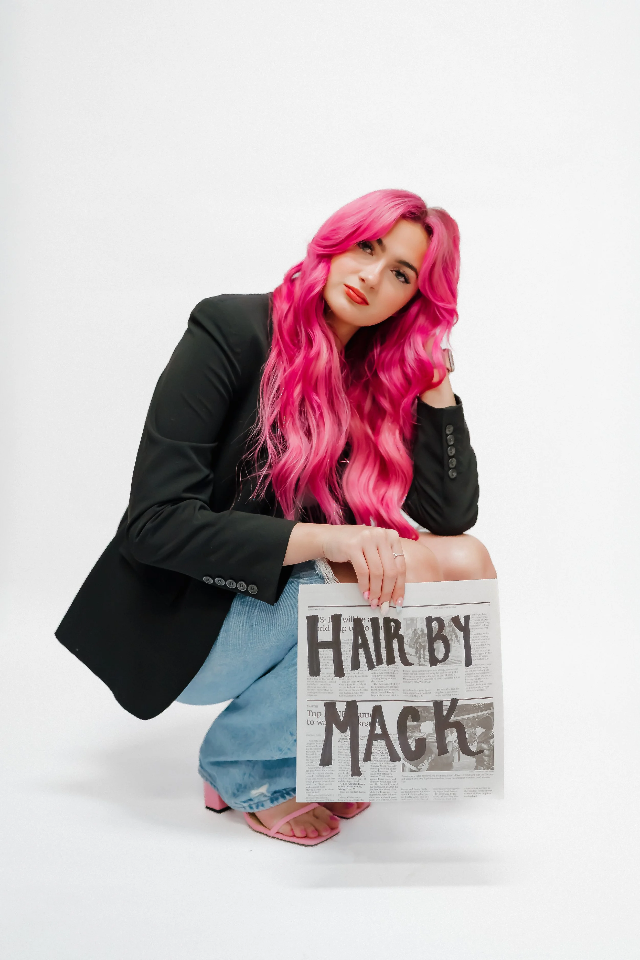 Woman with long pink hair wearing a black blazer and light blue denim skirt, holding a newspaper with the headline 'HAIR BY MACK', standing against a plain white background.