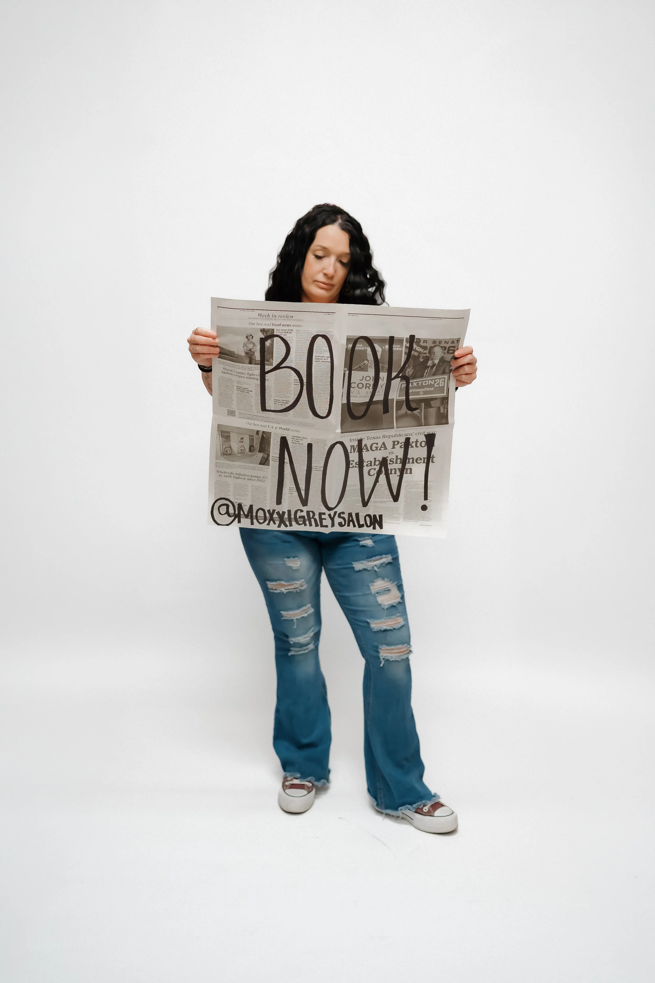 Woman with dark curly hair wearing ripped jeans, white sneakers with red accents, holding a newspaper with "BOOK NOW!" written across it in bold letters, standing against a plain white background.