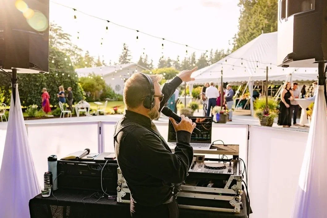 DJ with headphones and microphone playing music at an outdoor wedding reception, with guests socializing under tents and string lights in the background.