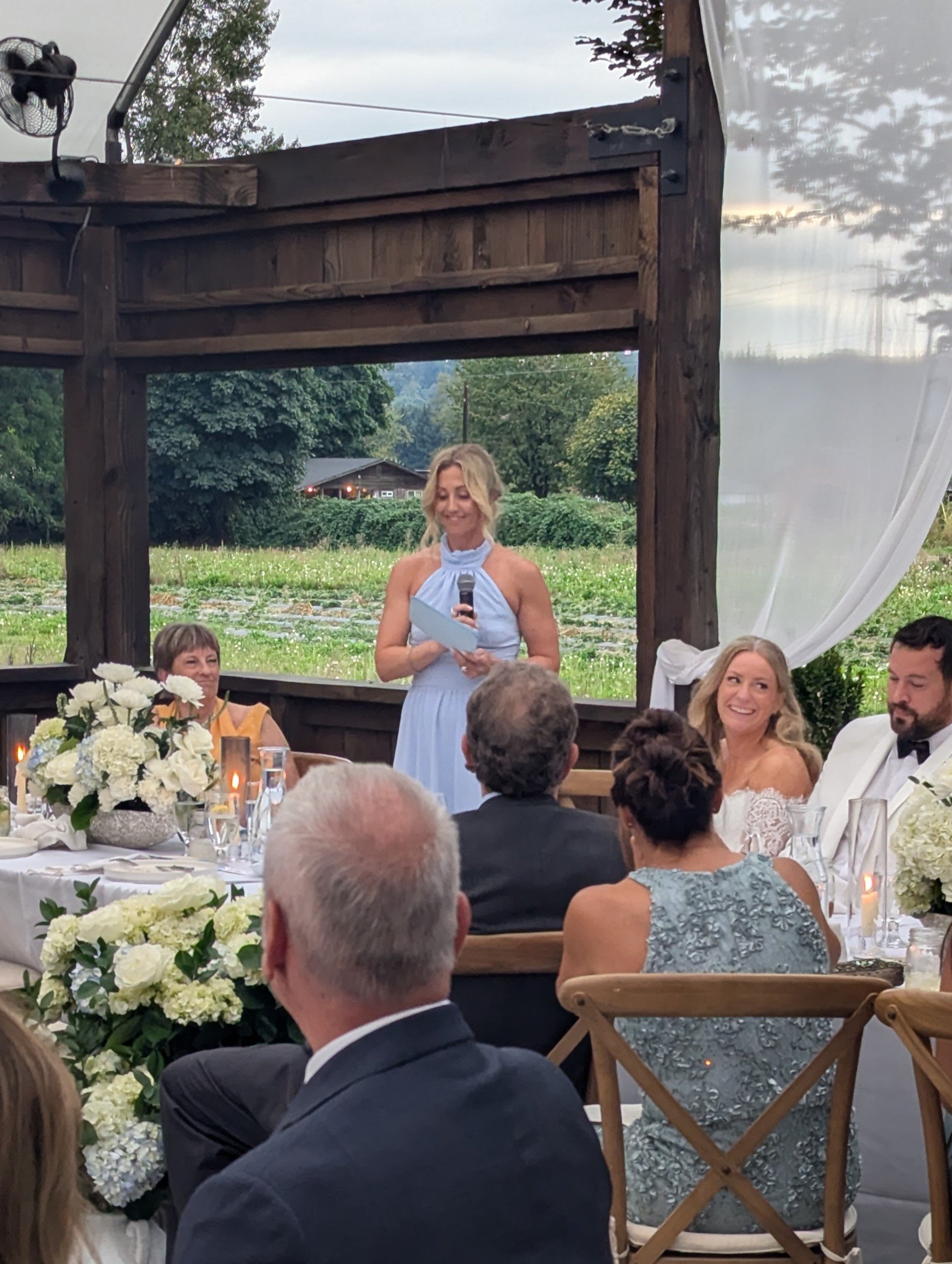 A woman in a light blue dress giving a speech at a wedding reception, standing behind a table with floral centerpieces, with guests seated in front of her, in an outdoor rustic setting with greenery and a wooden structure.