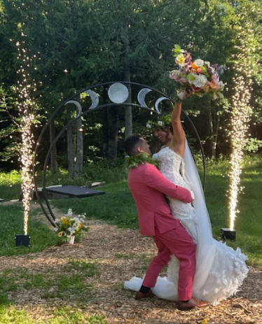 A wedding couple kissing outdoors beneath a moon phase arch, with one raising a bouquet and sparklers in the background.