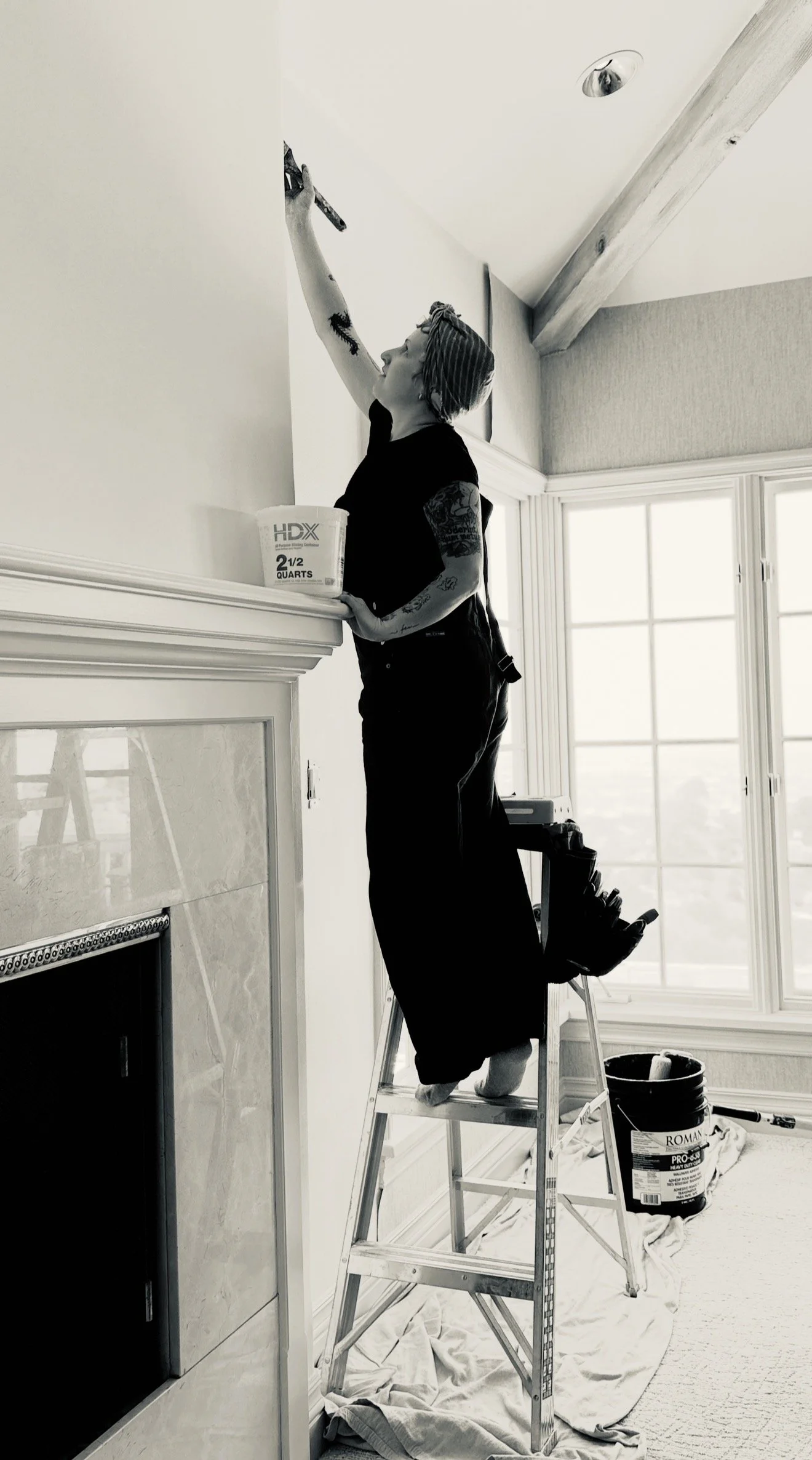 A person standing on a step ladder painting a wall near a ceiling with a roller brush, a bucket of paint on the mantel, and large windows in the background.