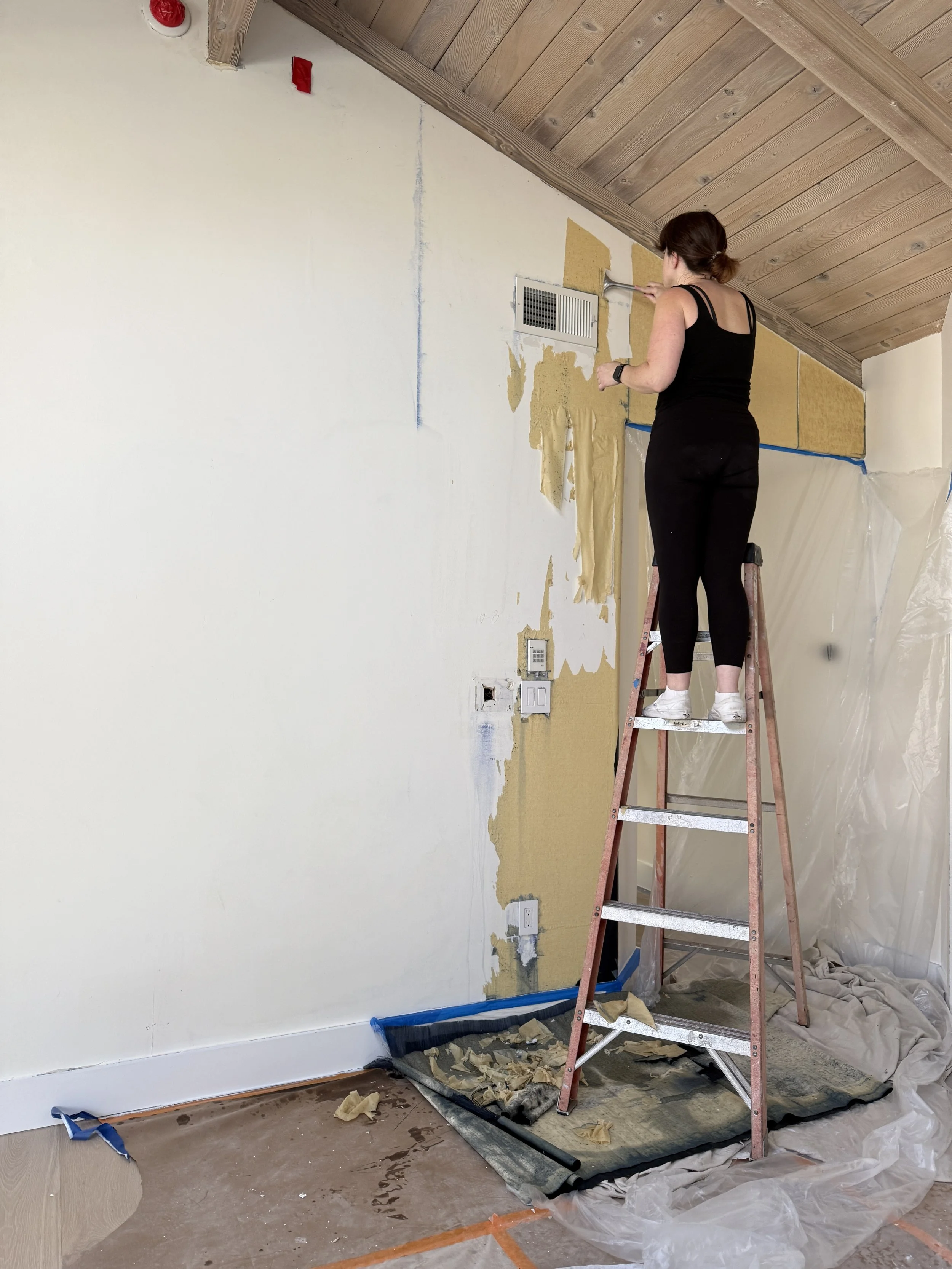 A woman standing on a step ladder, scraping paint or wallpaper from a wall in a room under renovation or painting, with construction tools and plastic sheeting on the floor.