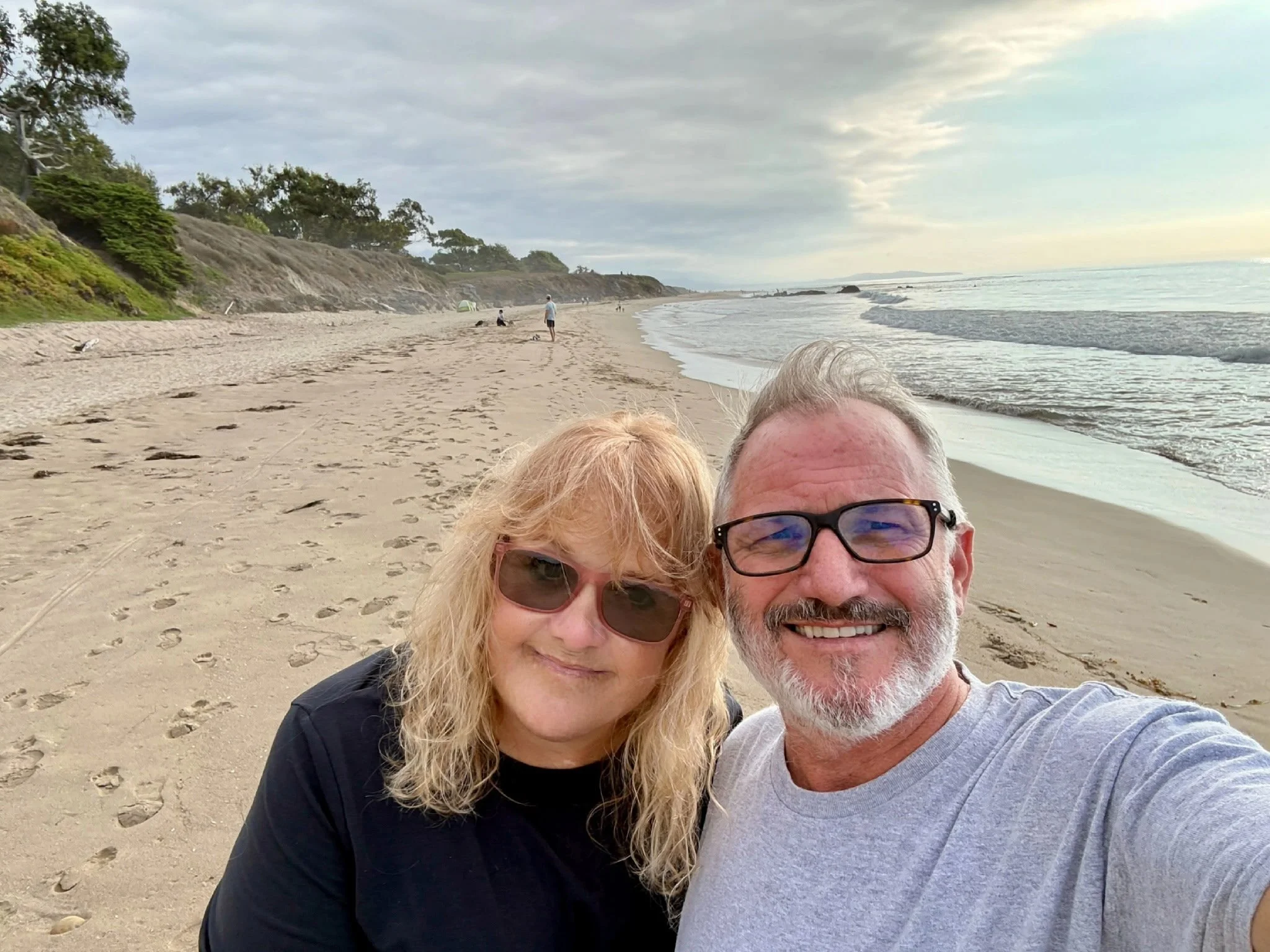 A smiling couple takes a selfie on a beach with footprints in the sand, facing the camera, while other people walk or sit in the background near the shoreline under a cloudy sky.