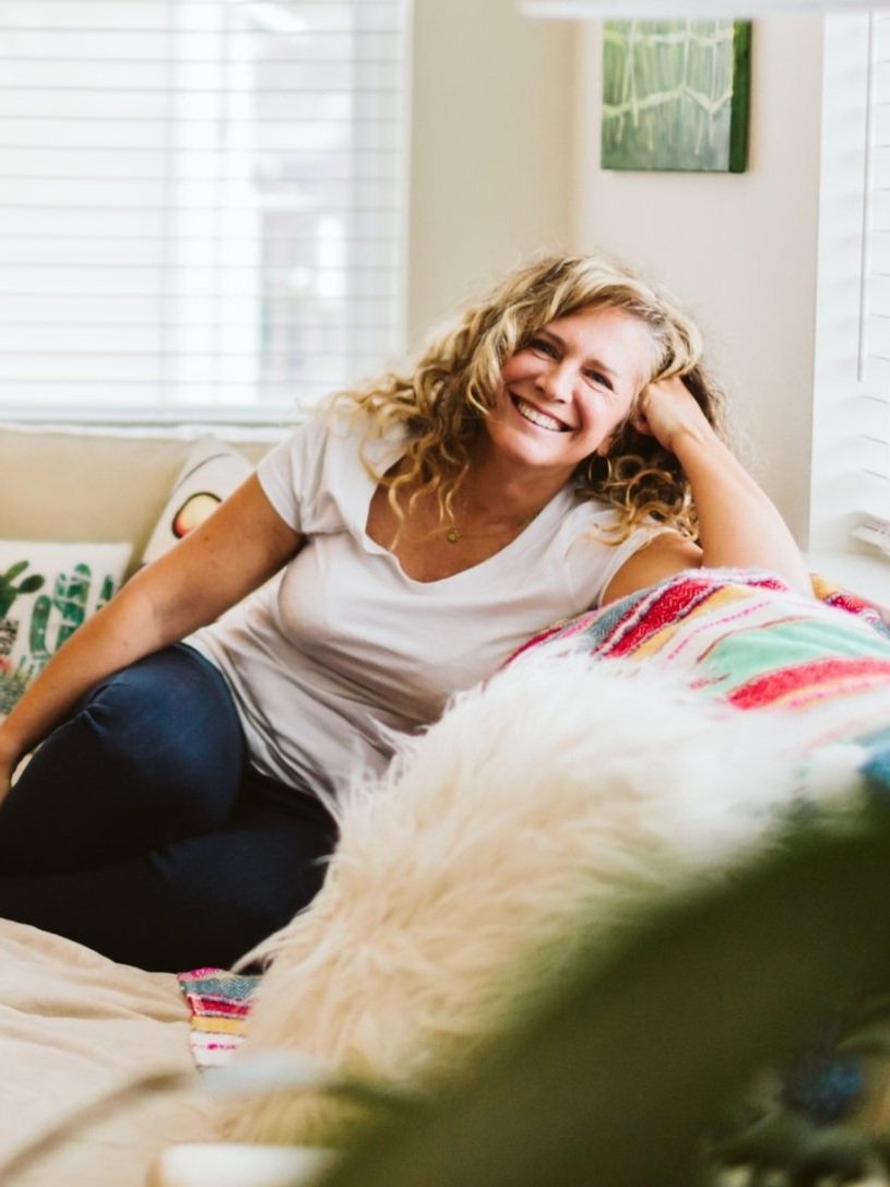A woman with curly blonde hair smiling, sitting on a couch in a bright room with white window blinds and a green painting on the wall, next to a fluffy white dog.