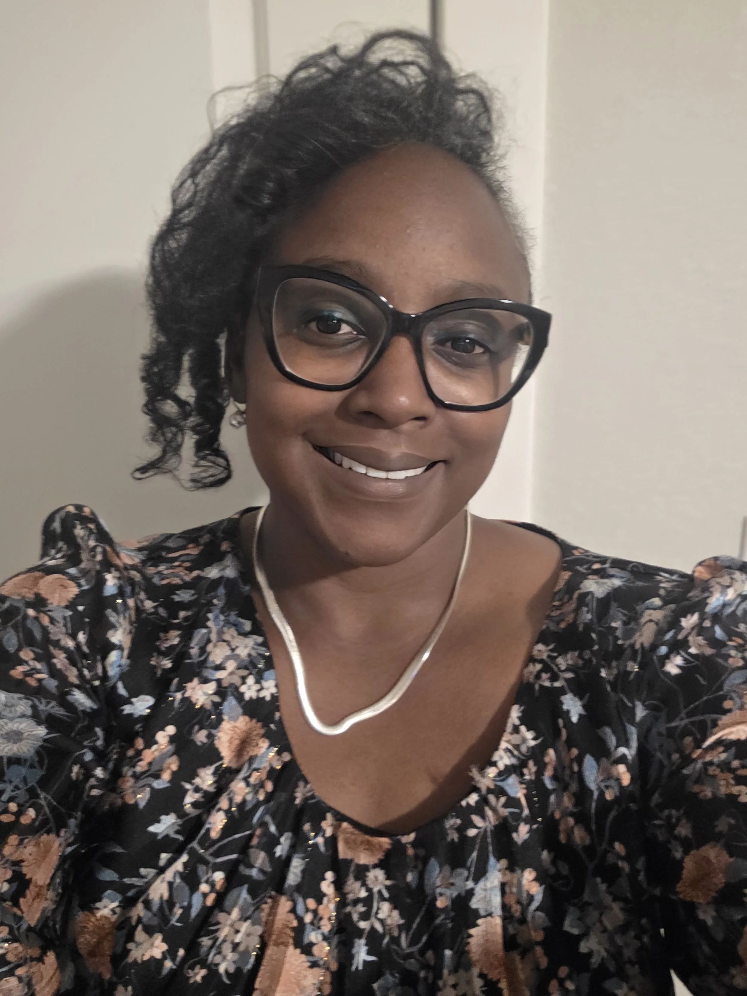 A woman with glasses, short curly hair, and wearing a floral dress and a necklace, smiling at the camera.