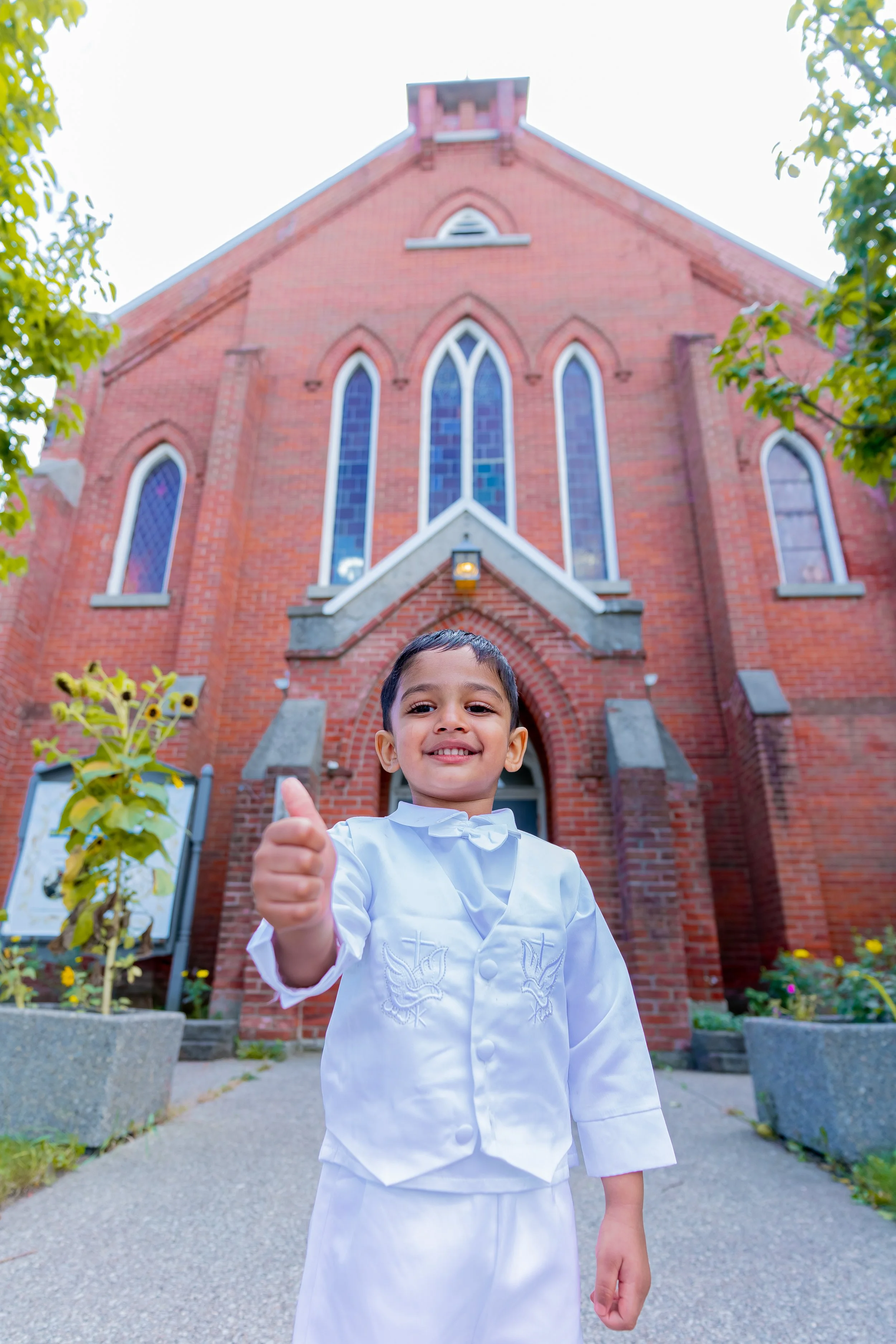 A young boy dressed in a white outfit, smiling and giving a thumbs up in front of a large brick church with stained glass windows.