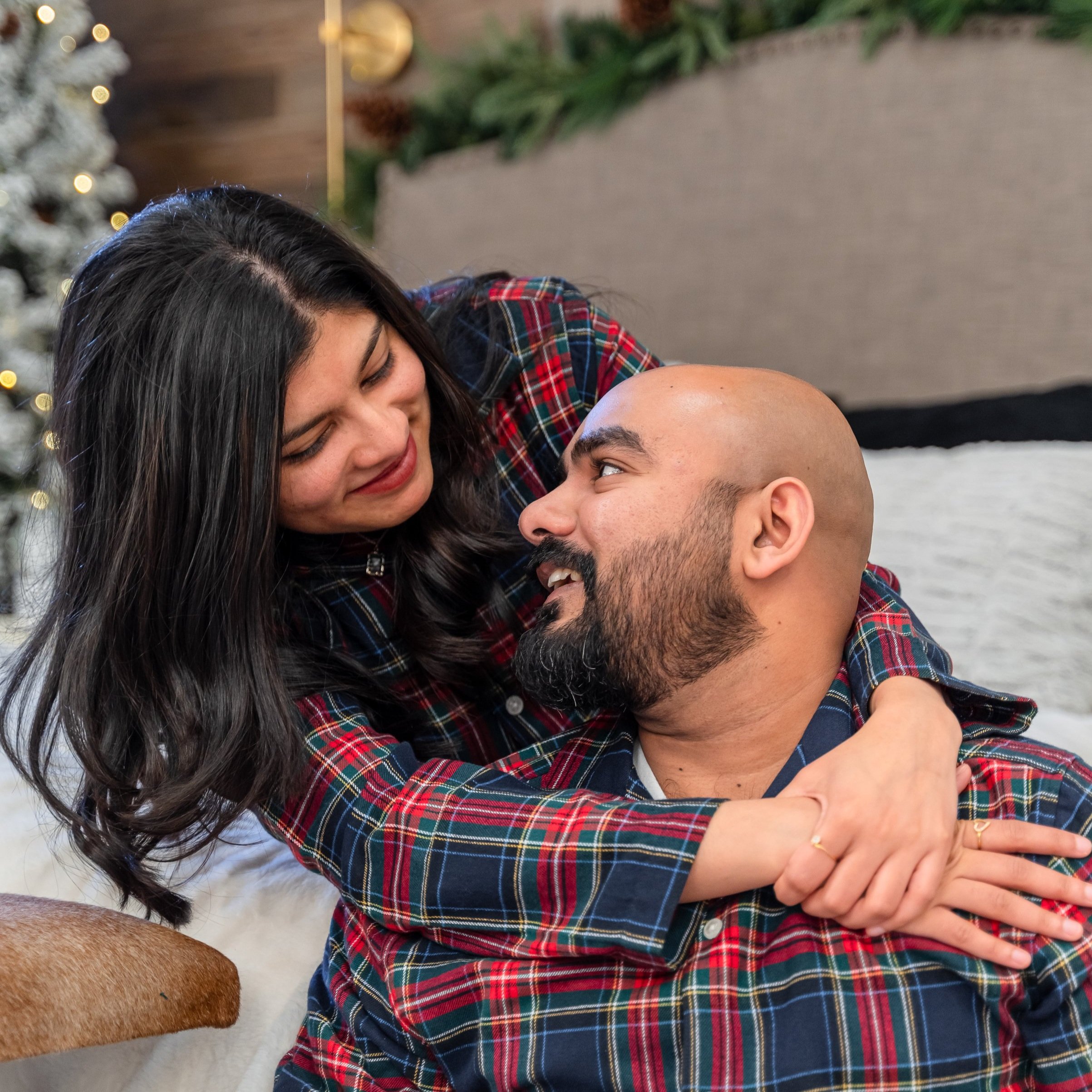 A woman and a man wearing matching plaid shirts share a warm embrace and look at each other lovingly in a room decorated for Christmas Photography Toronto, Michigan, New York & Texas, US