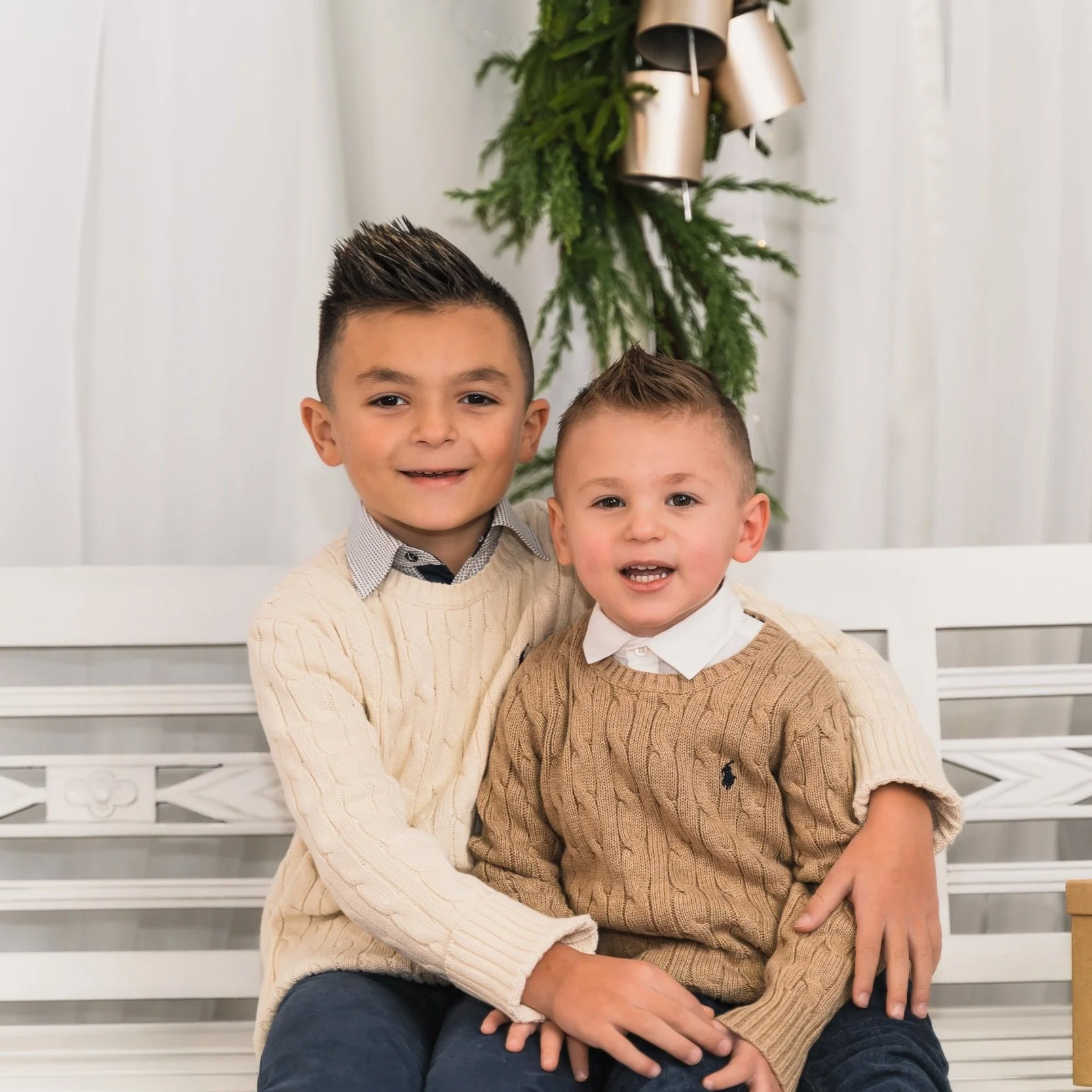 Two young boys sitting together in front of a Christmas tree, smiling at the camera. The older boy is wearing a cream sweater over a collared shirt, and the younger boy is in a tan sweater with a white collar shirt underneath.