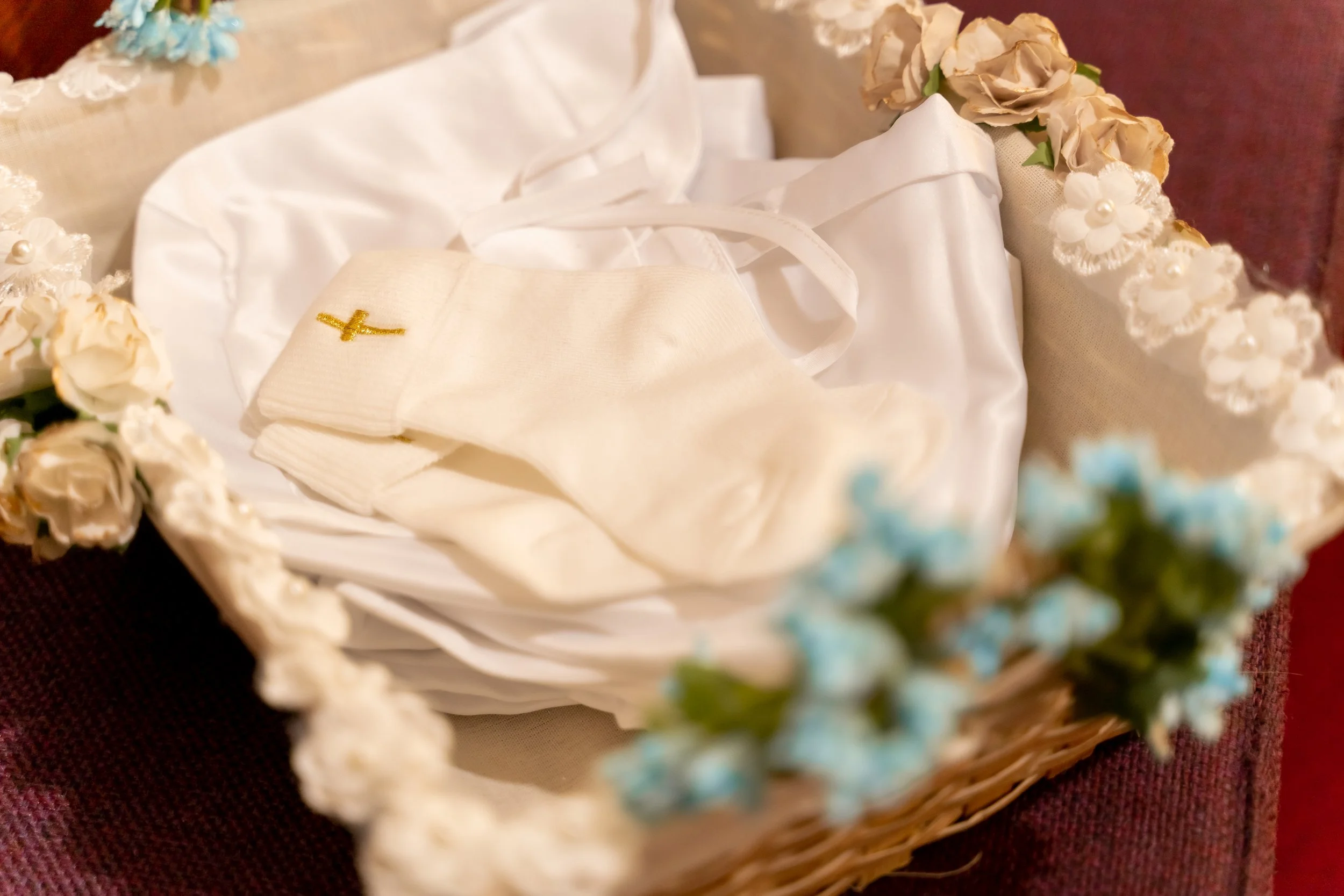 A wicker basket with white satin interior contains a white book, a cream-colored cloth with a small gold cross, and white linen with ribbons. The basket is decorated with artificial flowers in white, cream, and light blue, and is placed on a dark red fabric surface.
