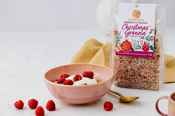 Superflora Christmas Granola in a bowl with yoghurt and raspberries scattered around and a pack of granola on the table too.