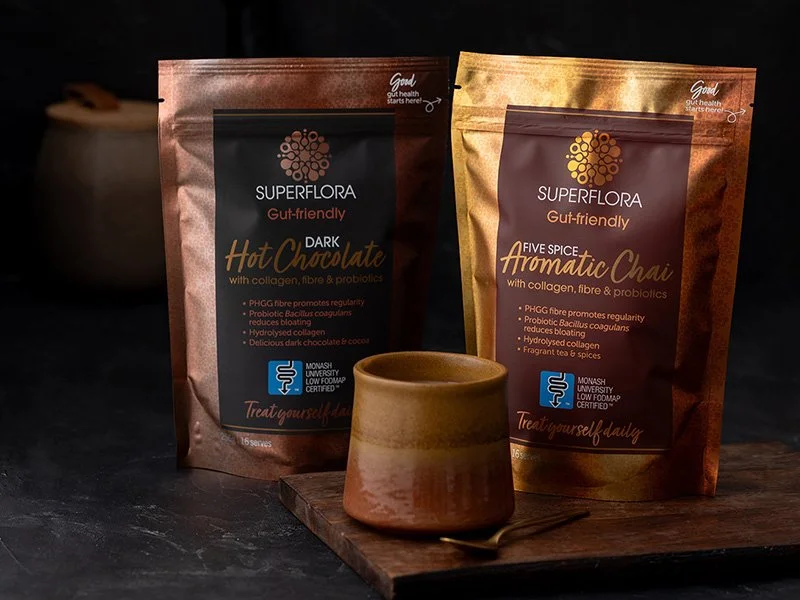 A packet of Superflora Dark Hot Chocolate, a pack of Gut-friendly Aromatic Chai and a cup of hot chocolate in a dark and moody kitchen.