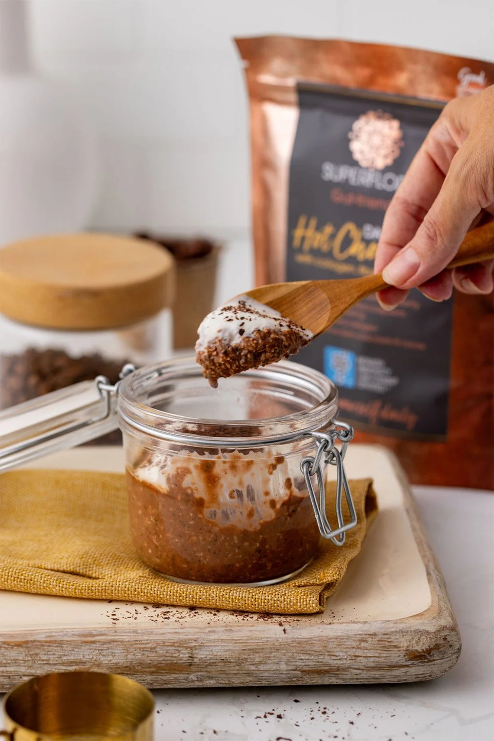 A pot of chocolatey overnight oats with a hand lifting a spoon with a good dollop of the oats, yoghurt and chocolate. A pack of Superflora Dark Hot Chocolate and container of coffee beans in the background.