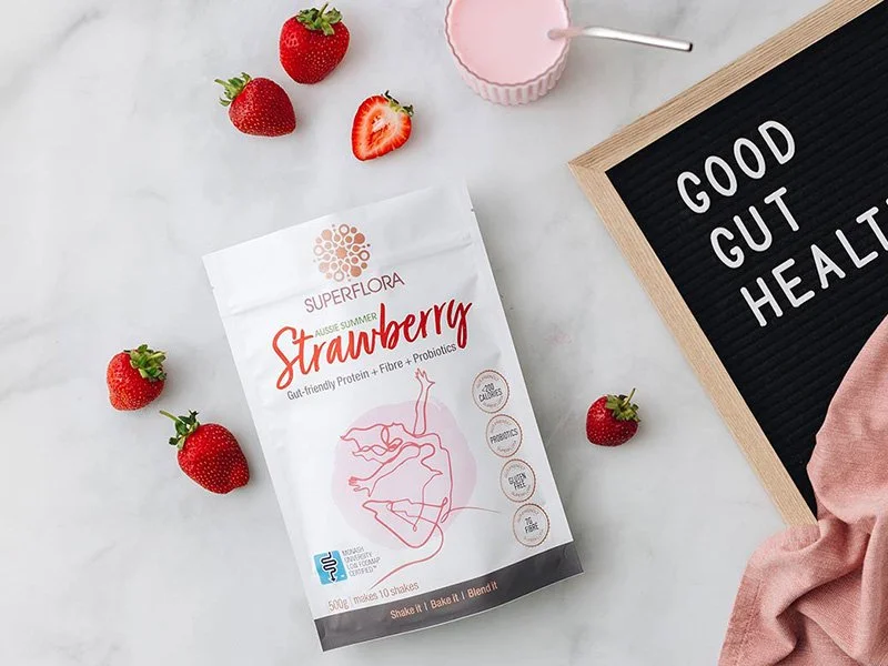 A pack of Superflora Aussies Summer Strawberry shakes along with a shake in a glass, strawberries and a pin board that says Good Gut Health Never Tasted so Good.
