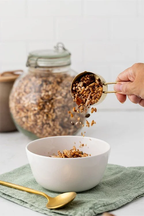 A hand pouring a cup of granola into a bowl.