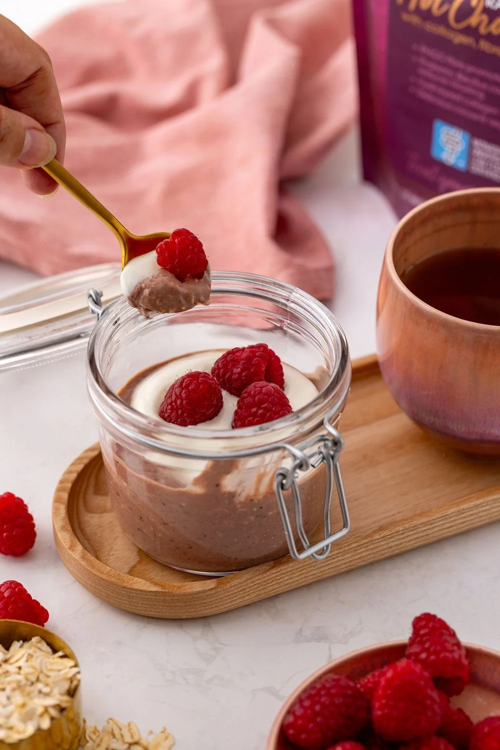 a hand scooping a spoonful of oavernight oats, lactose-free yoghurt and a raspberry out of a mason jar in a kitchen with a cup of coffee and a pack of Raspberry low FODMAP hot chocolate