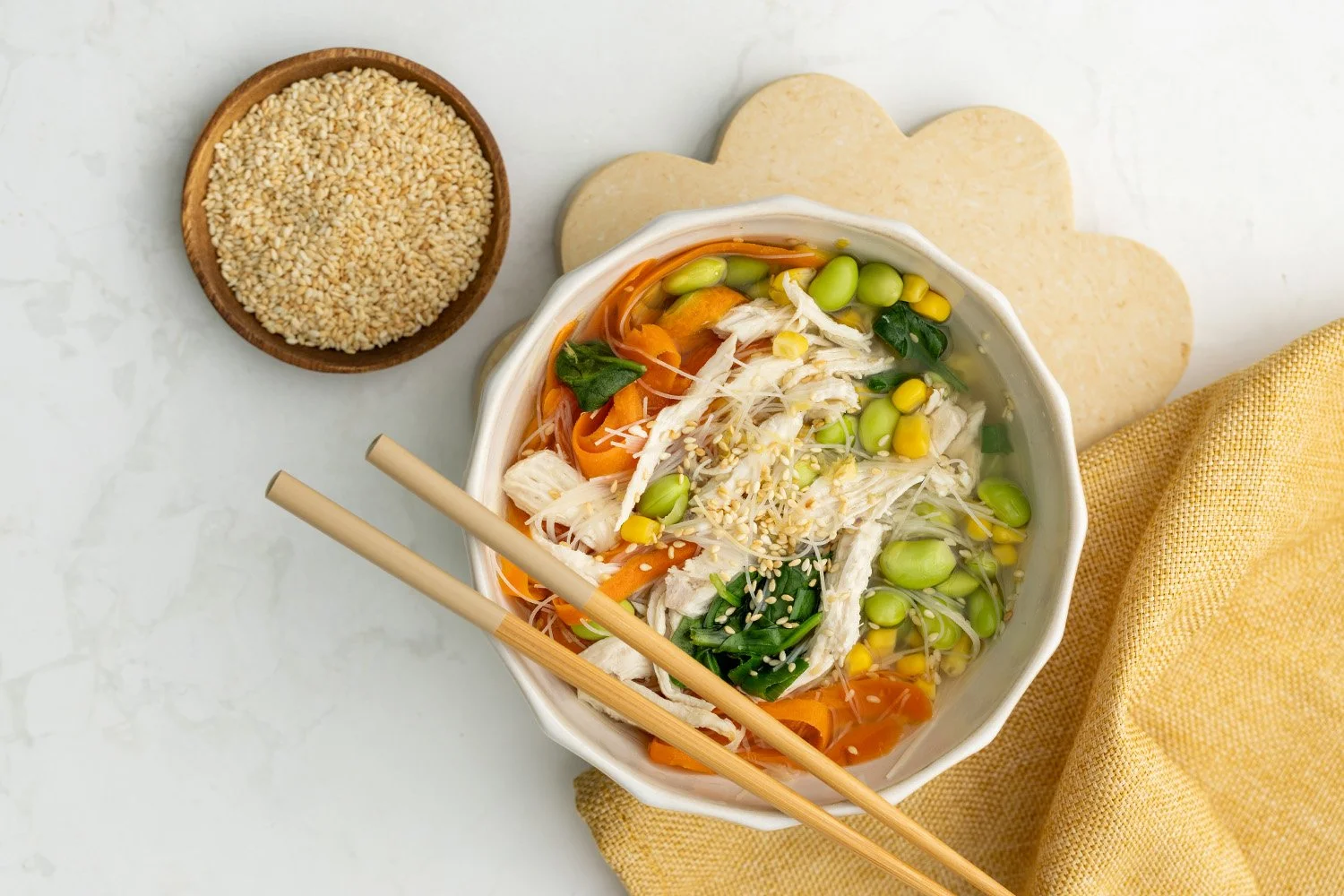 A bowl of noodles, chicken, bean and carrot sou =p with chopsticks and a bowl of sesame seeds.