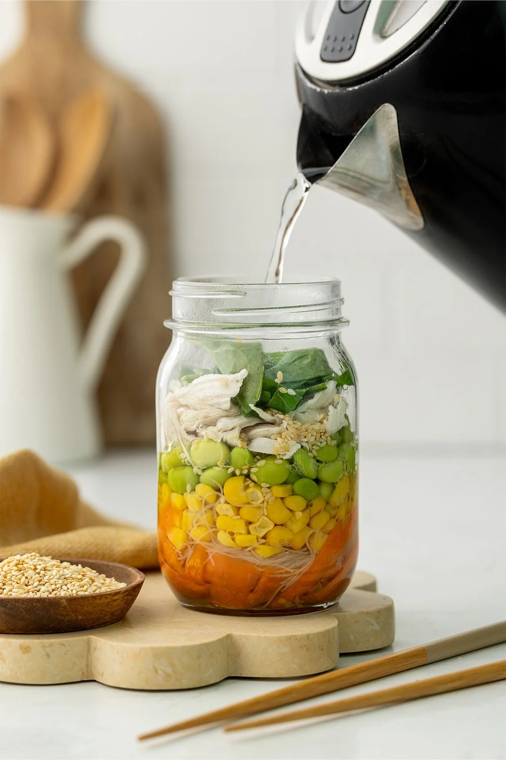 Water being poured from a kettle into a jar packed with colourful vegetables, noodles and chicken