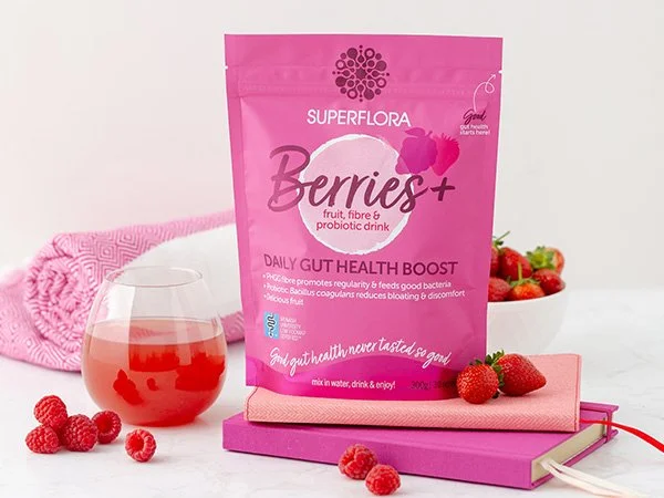 A packet of Superflora Berrie+ Daily Gut Health Boost with a glass of boost, some rasoberries a towel and a book.