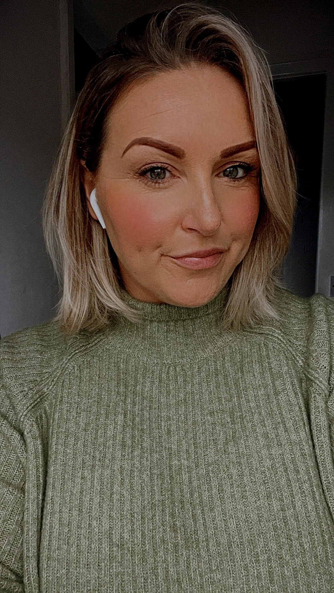 A woman with blonde hair, wearing a green ribbed turtleneck sweater, and white wireless earbuds in her ears, smiling softly at the camera.
