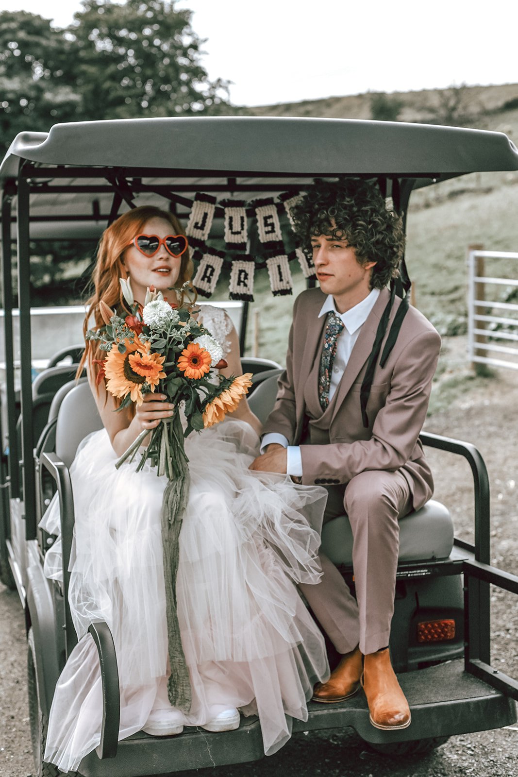 a cute bride wearing heart shaped sunglasses on the back of a gold buggy with her curly haired groom