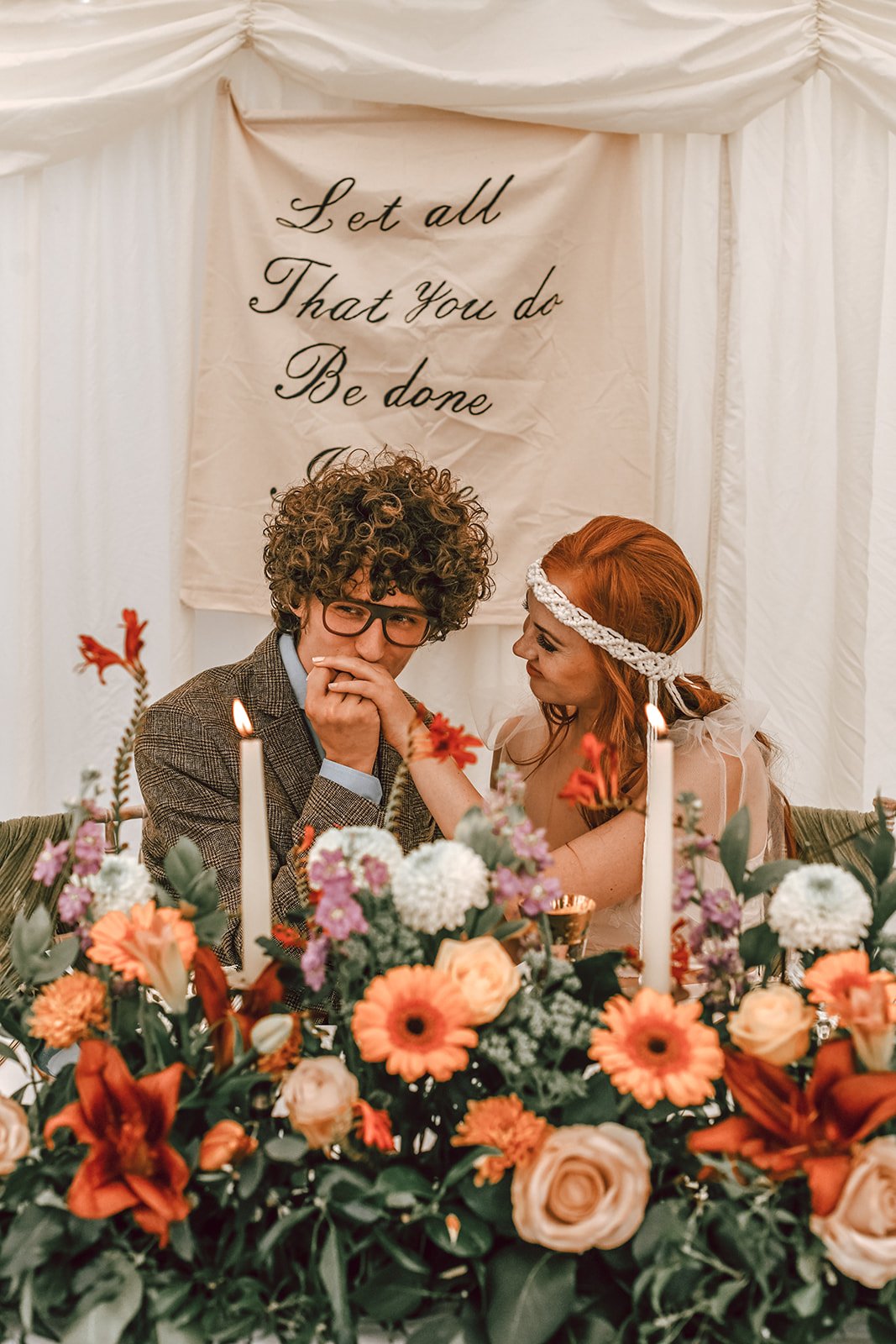 a bride and groom sat at their sweetheart table with flowers in the foreground and a banner in the background 