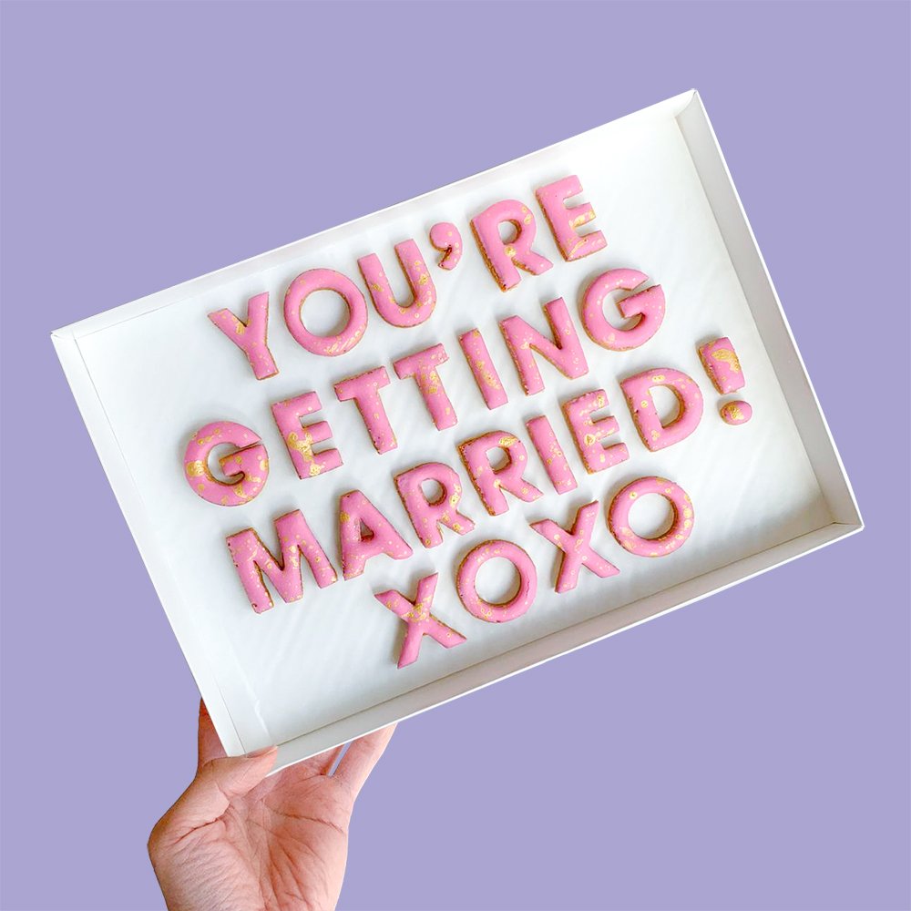  A white gift box of pink customised cookies that spell the wordS YOU’RE GETTING MARRIED! XOXO 