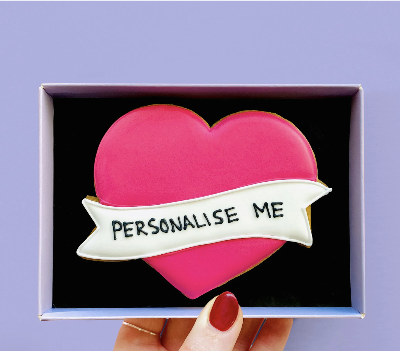  A bright pink heart-shaped cookie in a box that has a white customisable banner through it with the words ‘personalise me’ in black icing. 