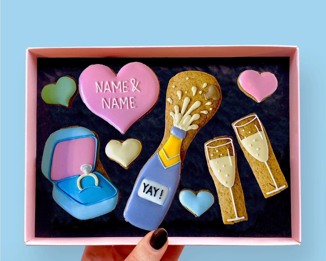  A cute engagement gift box of pastel coloured cookies in a range of shapes including 2 glasses of champagne, a bottle of champagne that says ‘yay’. 4 small hearts, and one big heart that can be customised with the names of the happy couple. 