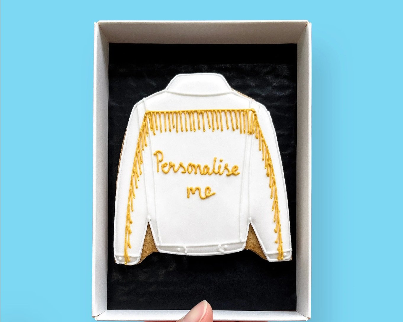  In a white gift box with a black inlay there is a white iced jacket with PERSONALISE ME iced in yellow with additional iced yellow fringing on the back of the jacket. 