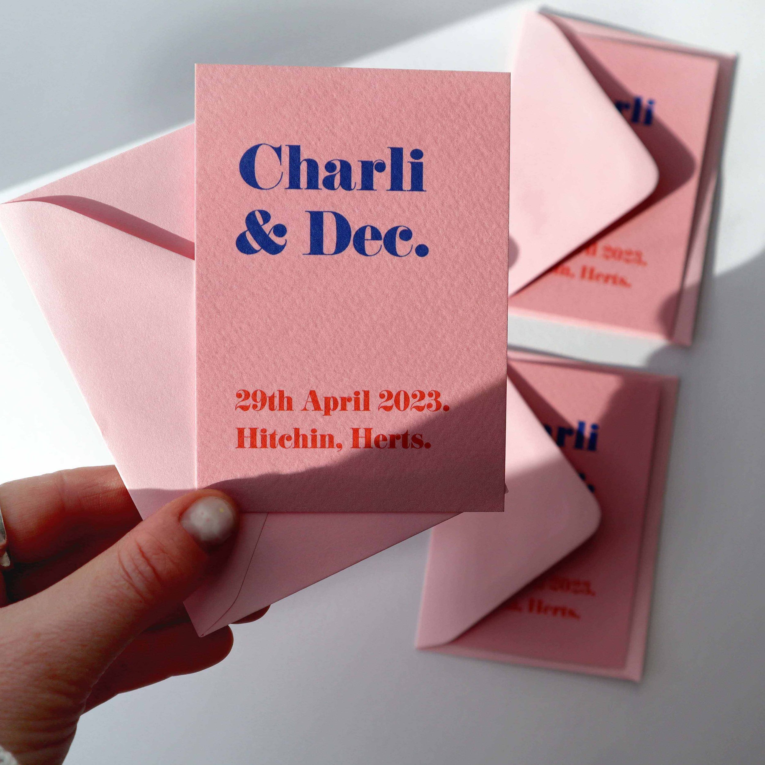 A hand holding up a contemporary wedding invite that’s millennial pink with the couple’s names in bold blue type and the wedding date in red. 