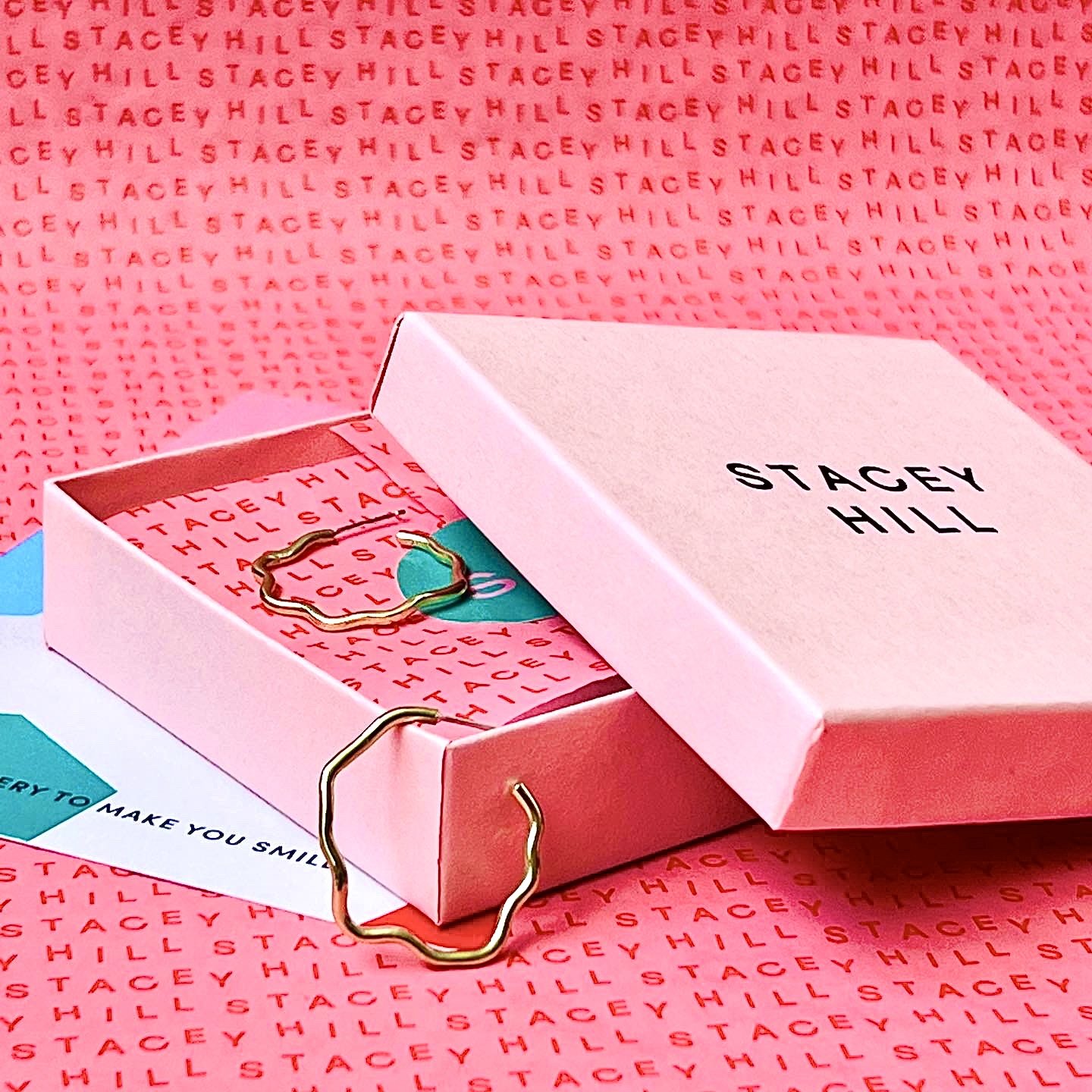  A pink Stacey Hill jewellery box with a pair of gold wiggly earrings inside. 