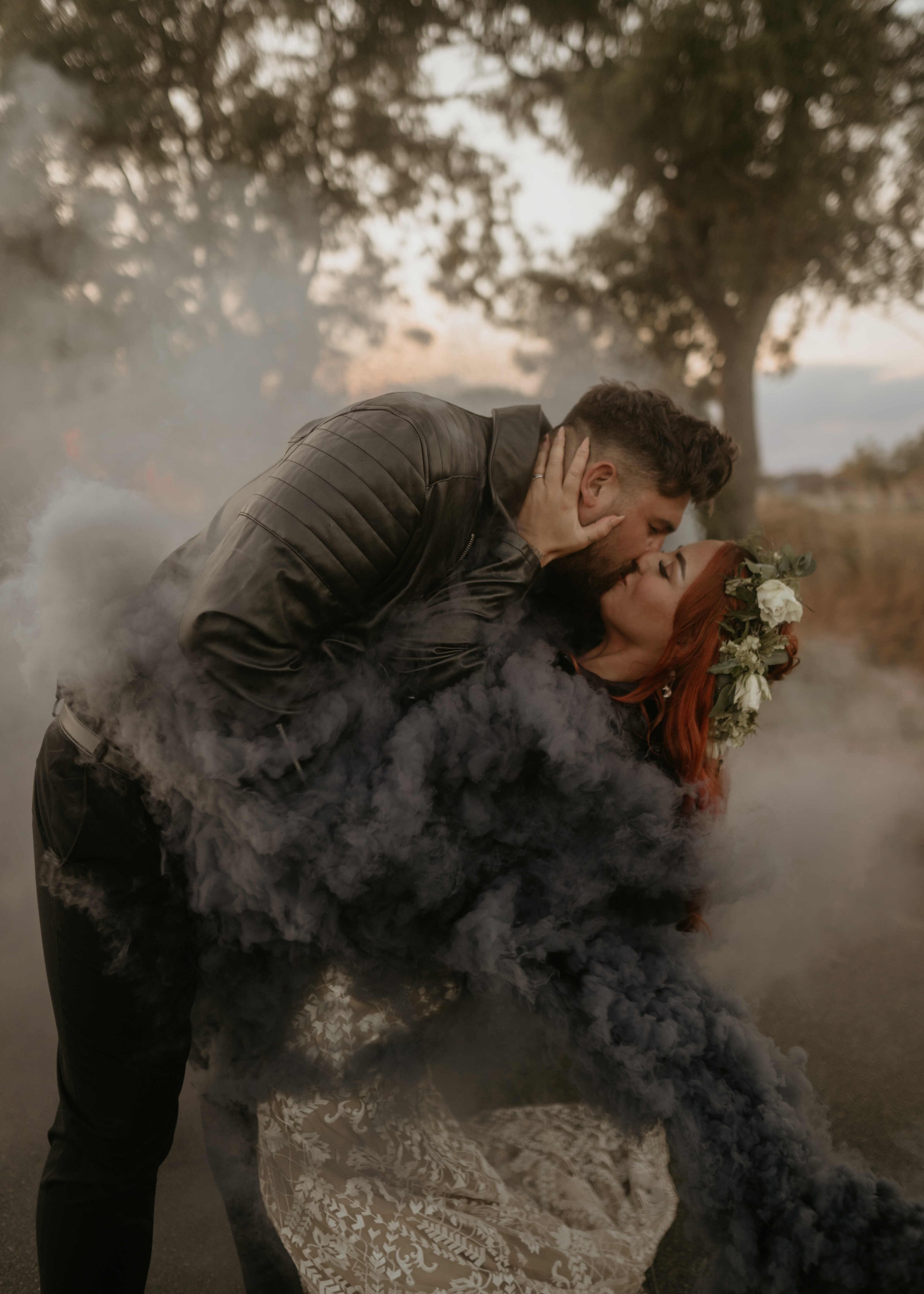  An unconventional couple kissing on their wedding day.They are both wearing leather jackets and you can see them trough a grey cloud of smoke. 