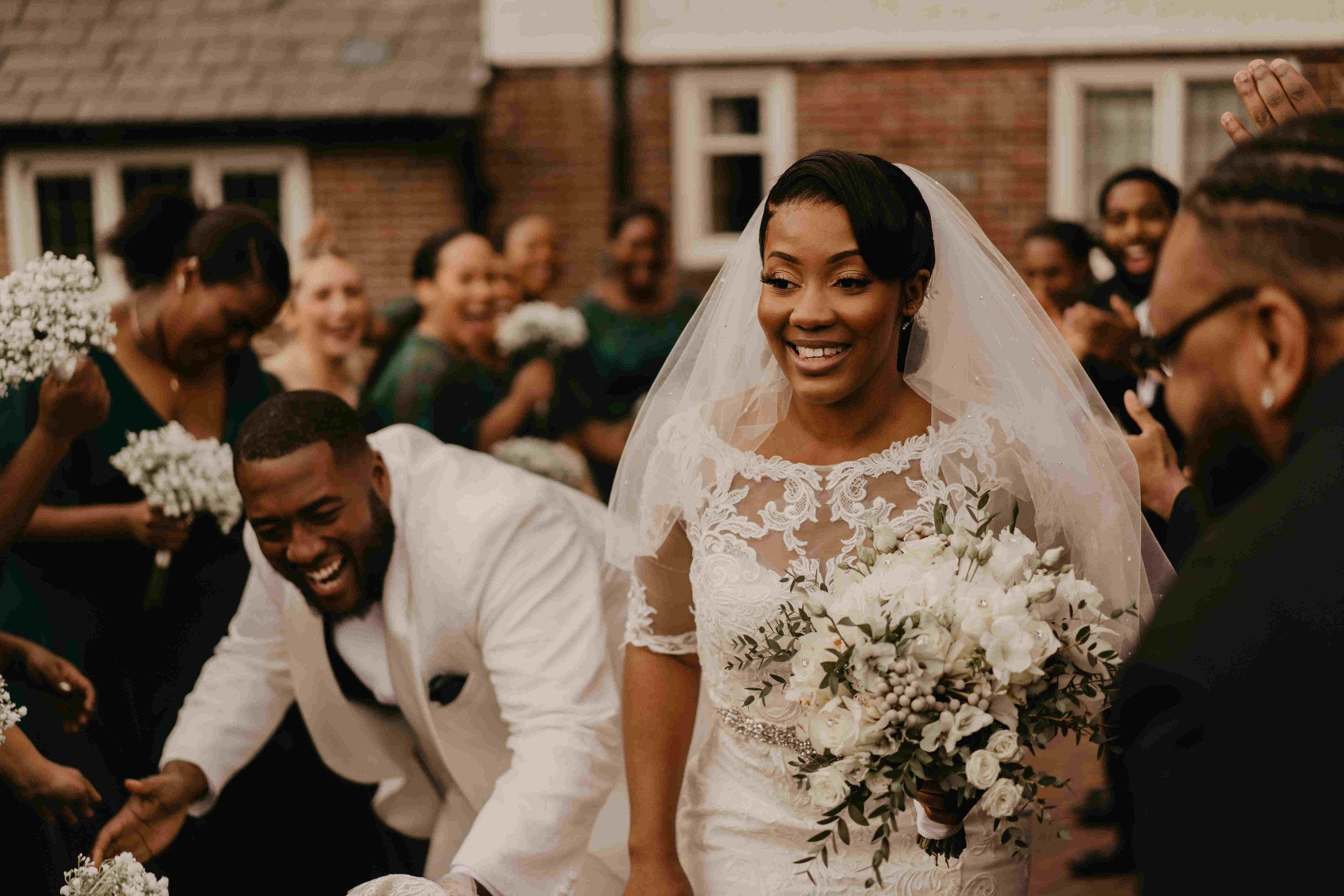  A Black bride and groom looking so happy on their wedding day. The bride has a big beautiful smile and is wearing a veil and a lace dress. The groom is wearing a white jacket and is crouching down slightly. 