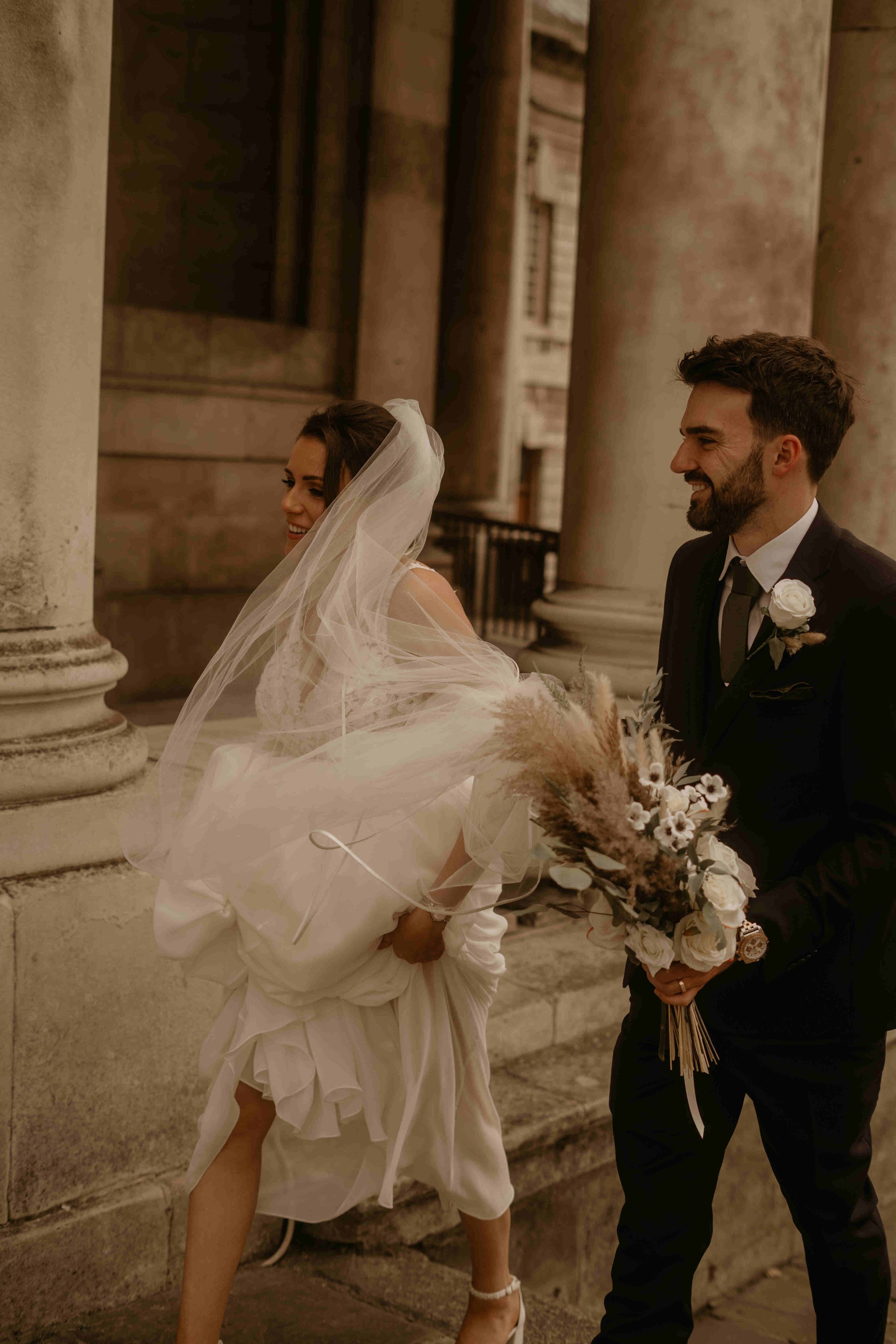  A bride and groom mid-stride. The bride is wearing a long veil  and holding up the length of her dress. 