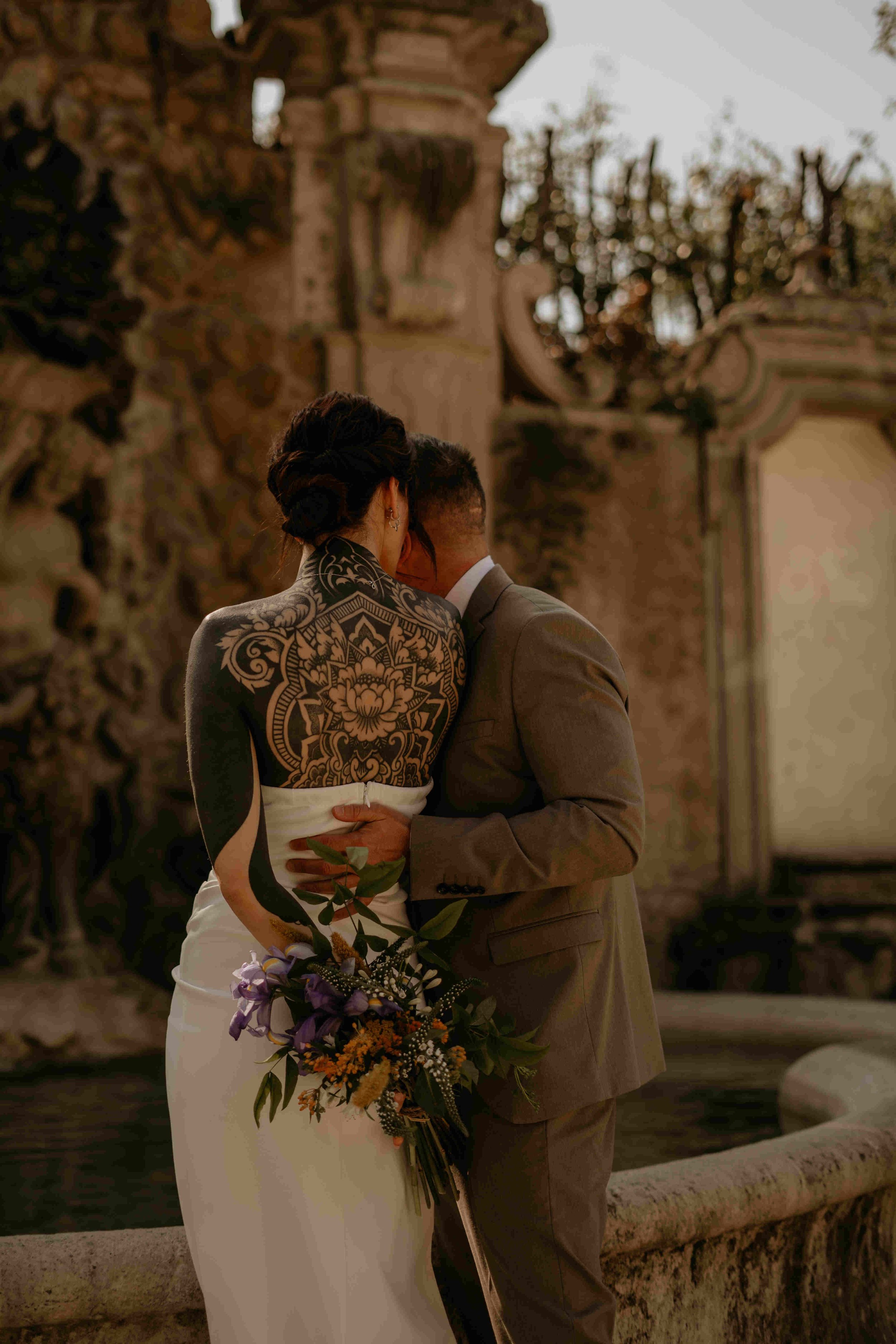  A photo of a bride and groom with their backs to the camera in a sunny destination. The bride has a low back dress and you can see her full back tattoo. 