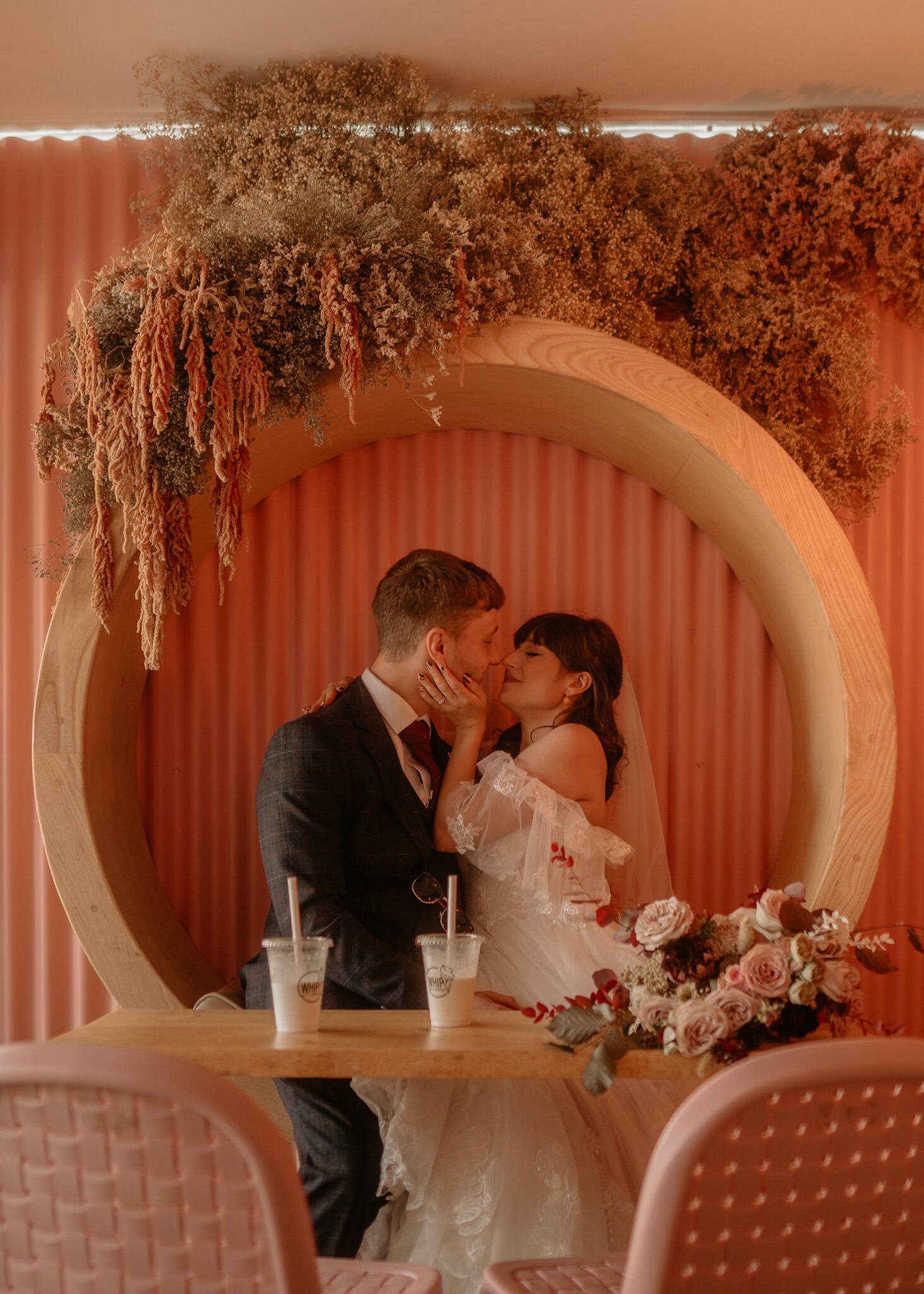  A bride and groom kissing inside a circular wooden cove with a peach wall behind them. There are 2 milkshakes and a bouquet of flowers in front of them 