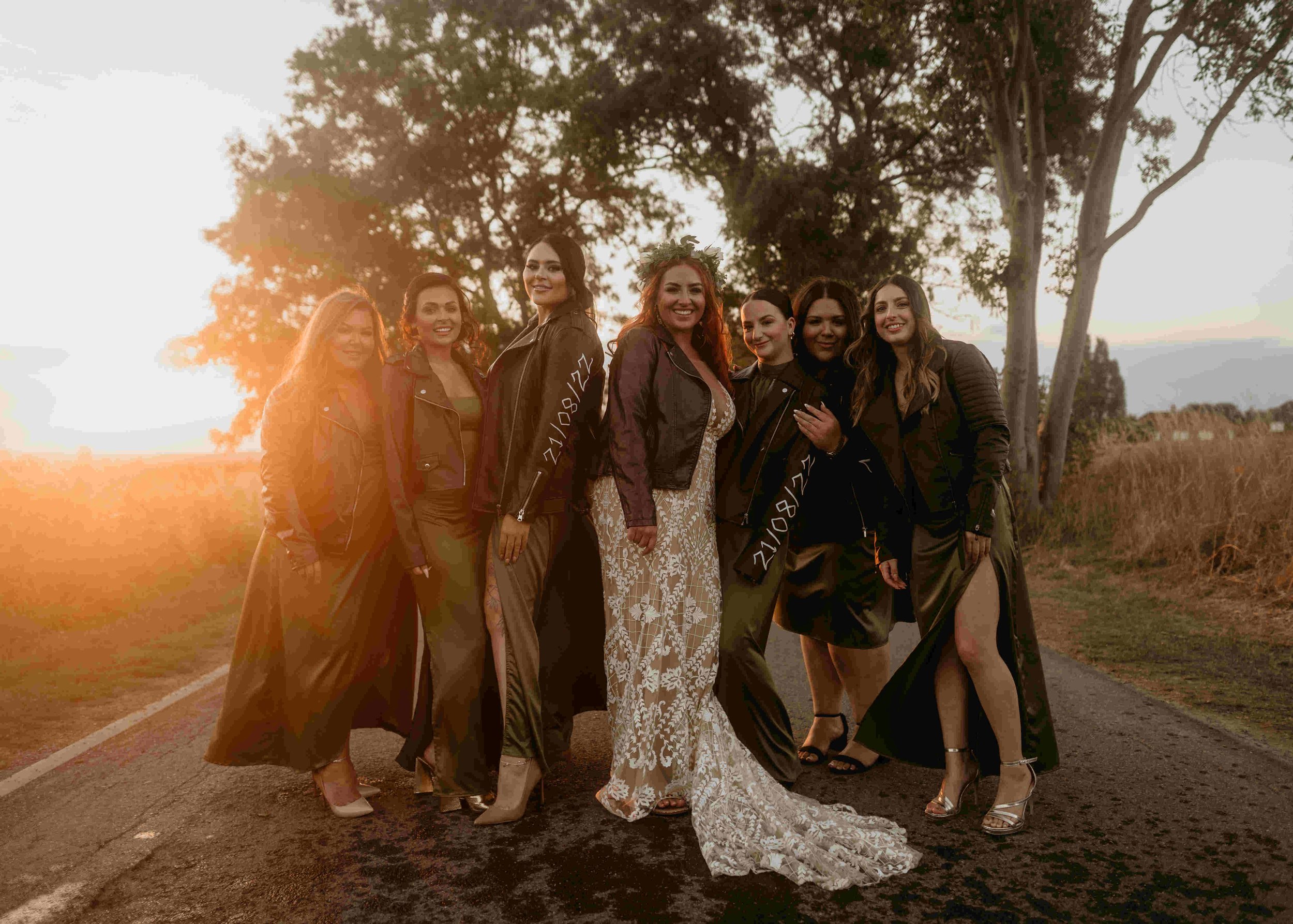  An alternative bride and her bridesmaids standing beneath a tree in the golden hour. They are all wearing leather biker jackets that have been customised with the wedding date. 