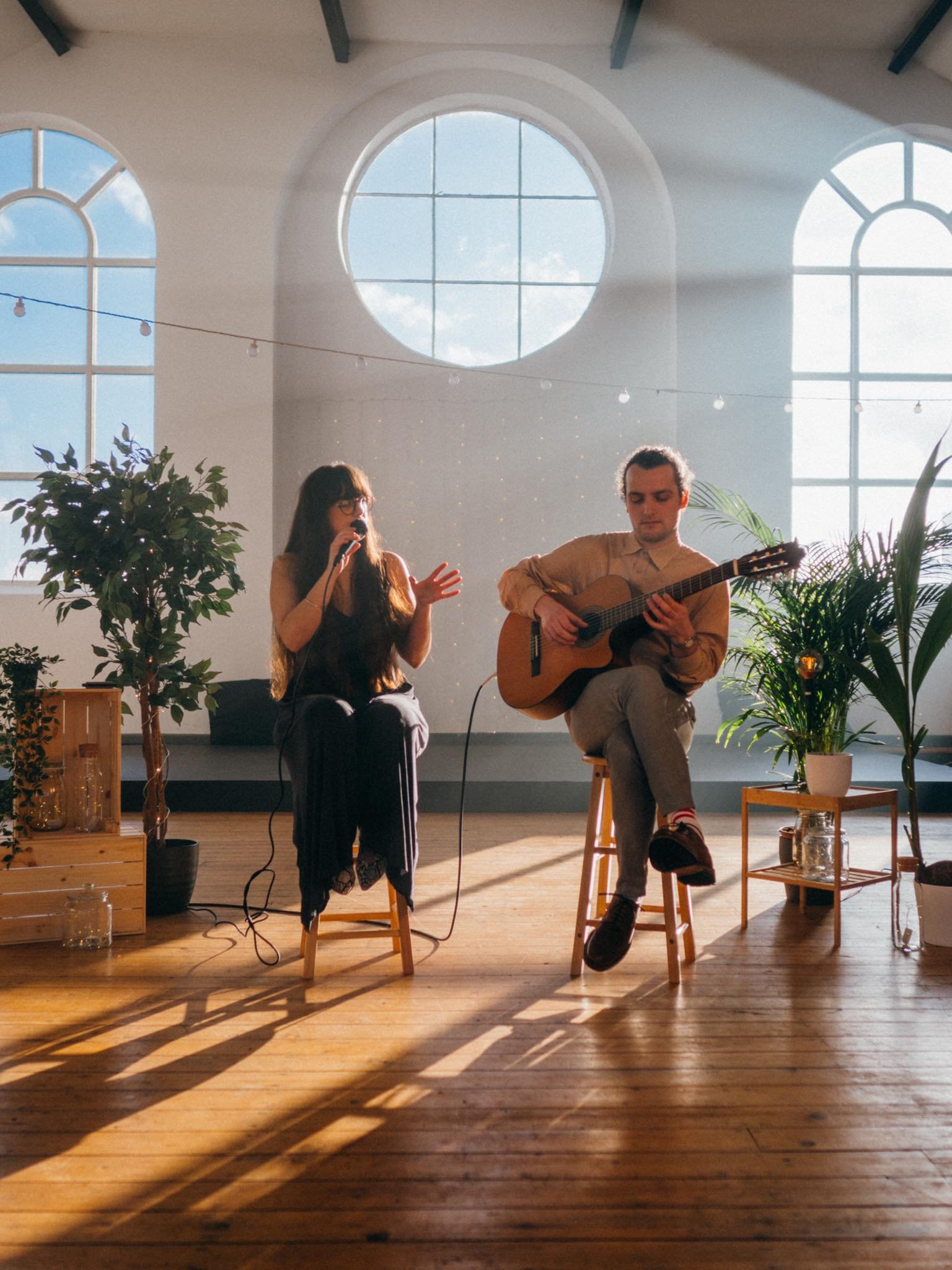  A male and female duet both sitting in wooden stools in a beautifully lit room with arched and circular windows. The woman is singing into a microphone and the man is playing a guitar. 