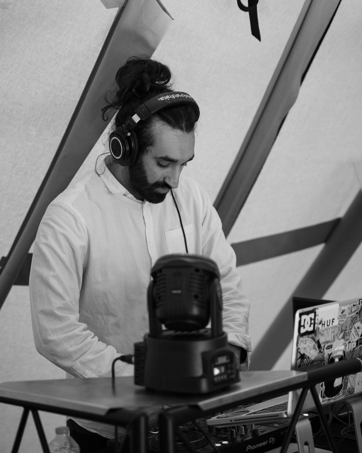  A black and white photo of a dark-haired man with his hair in a bun who is wearing headphones and appears to be djing. 