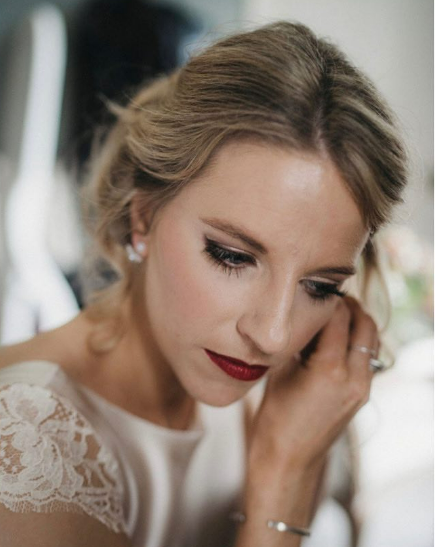  A photo of a bride putting her earring in. She’s wearing a full glam makeup with black eyeliner and a deep red lip. 