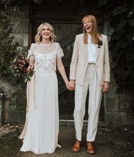  2 brides looking so happy on their wedding day. They are holding hands. One has mid-length blonde hair and is wearing a dress. The other has long red hair with a fringe and is wearing a cream suite. 
