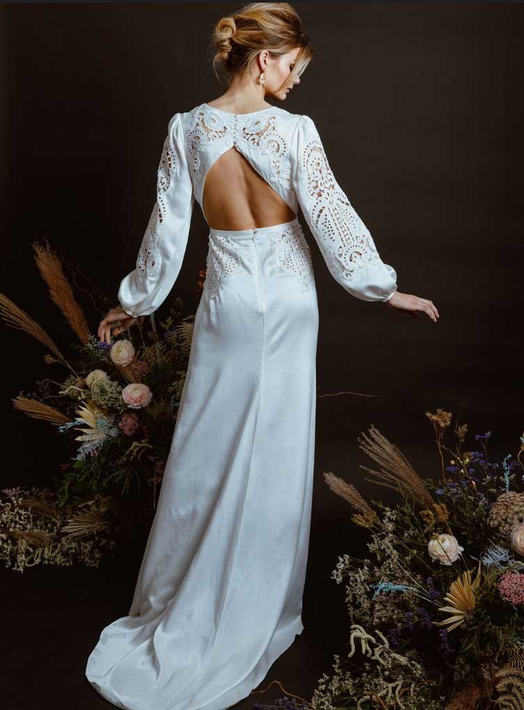  A bride standing with her back to the camera to showcase her backless dress. It has long sleeves and a beautiful lace detail. 