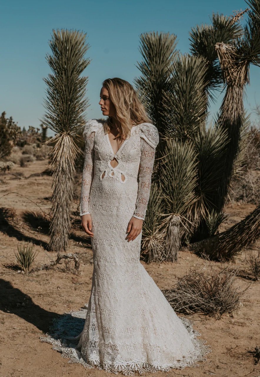  A blonde haired bride standing in the desert wearing a long-sleeved bohemian bridal dress. 